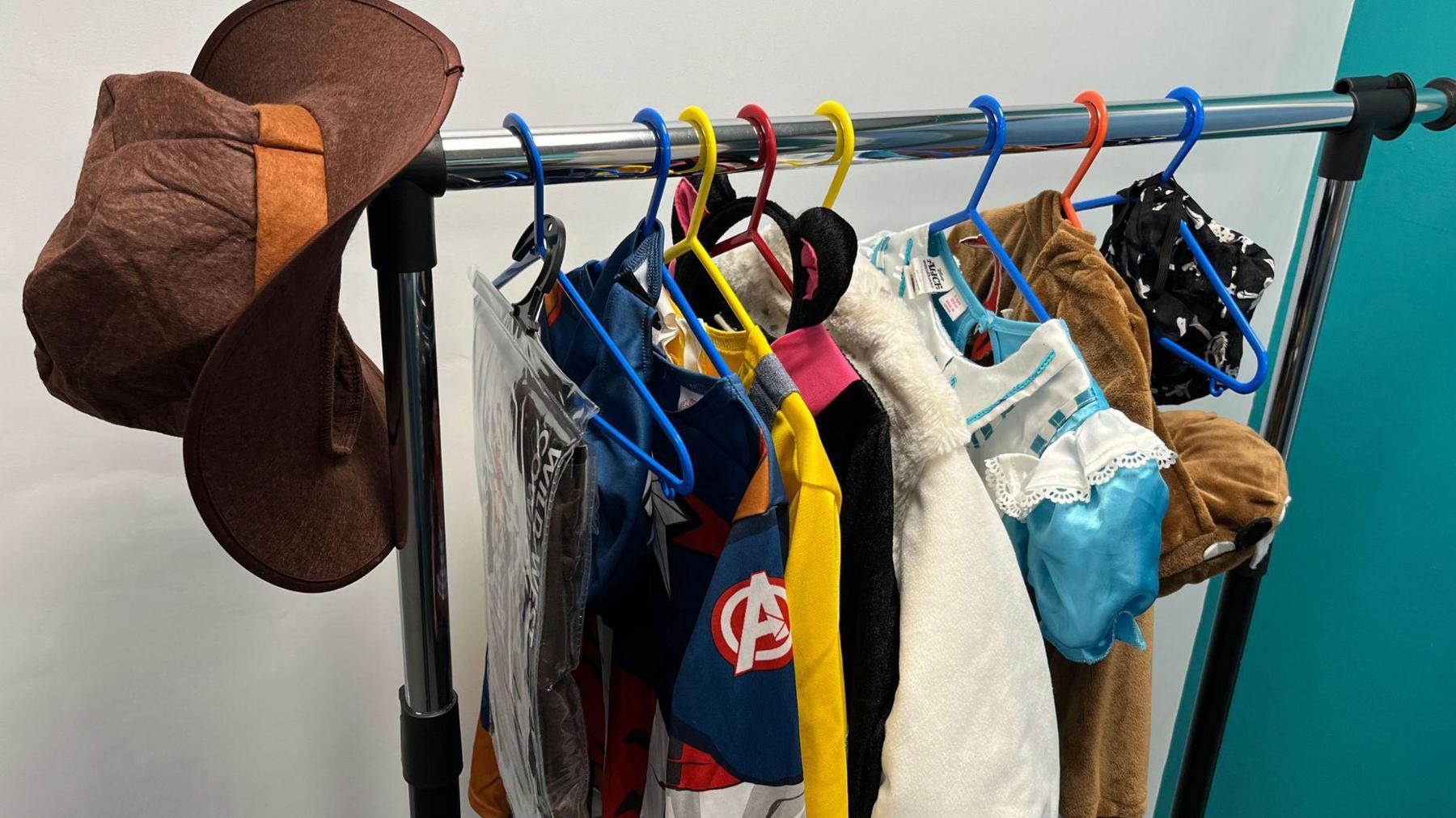 A metal clothes rail with several coloured hangers containing a selection of costumes. A brown cowboy hat is perched at one end.