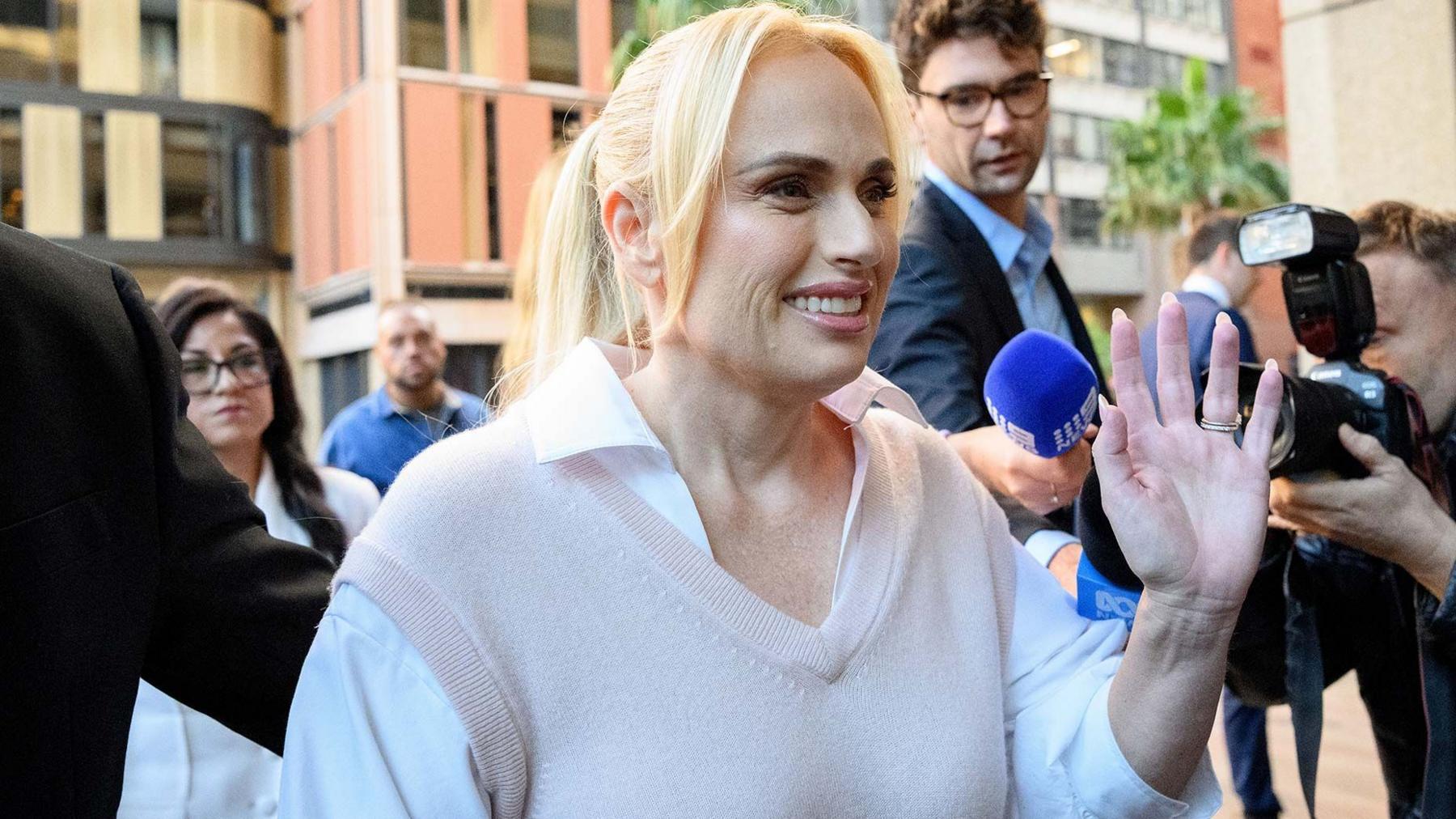 A woman waving at the camera outside court with blonde hair, wearing a white shirt and beige vest 