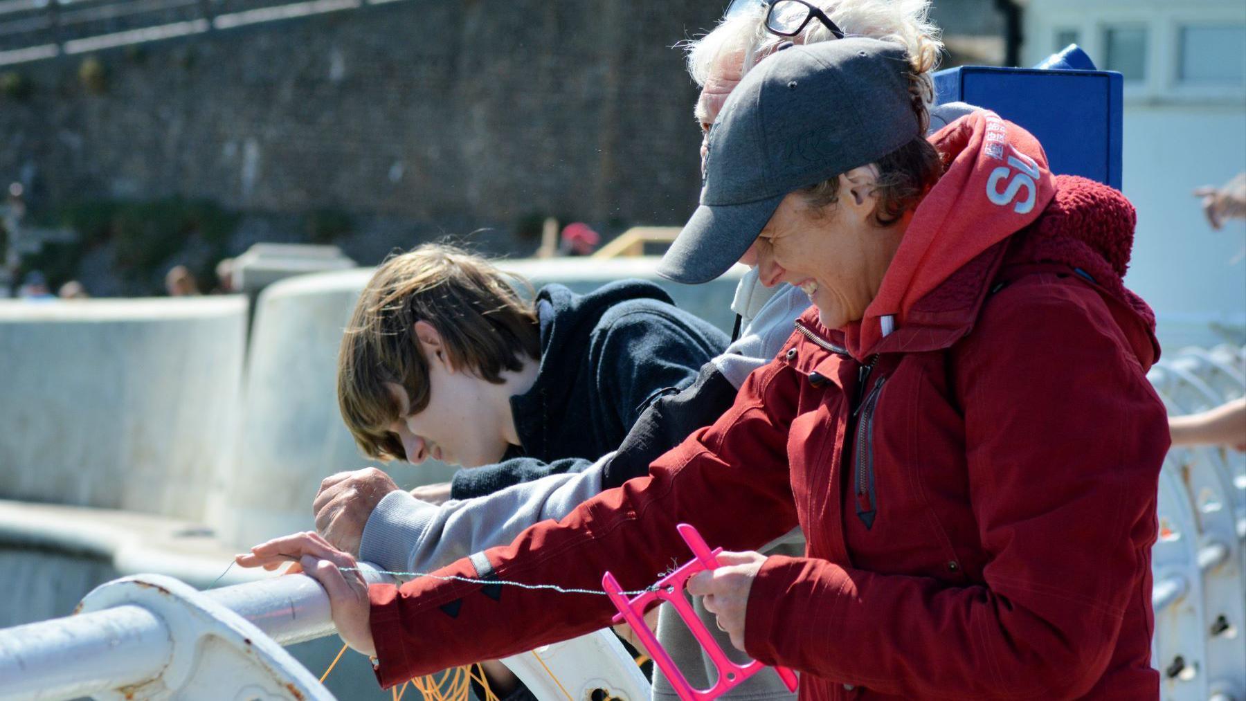 A woman and boy crabbing on Cromer Pier. The woman is wearing a red coat and blue cap, and is holding a pink crabbing line, while the boys has hair across his face and is looking over the edge of the pier