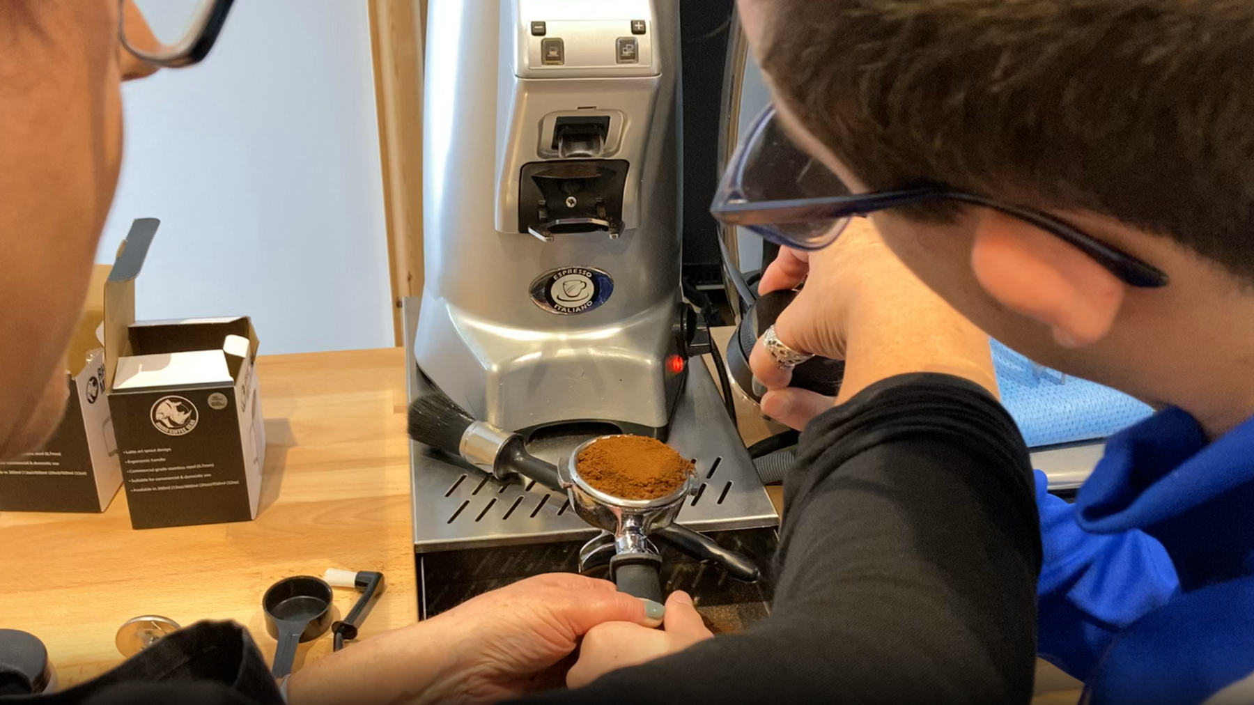The photo is taken from over the shoulders of a woman and a younger man as they operate a coffee machine. The woman is using her hands to guide the younger man's hands to use the machine. 