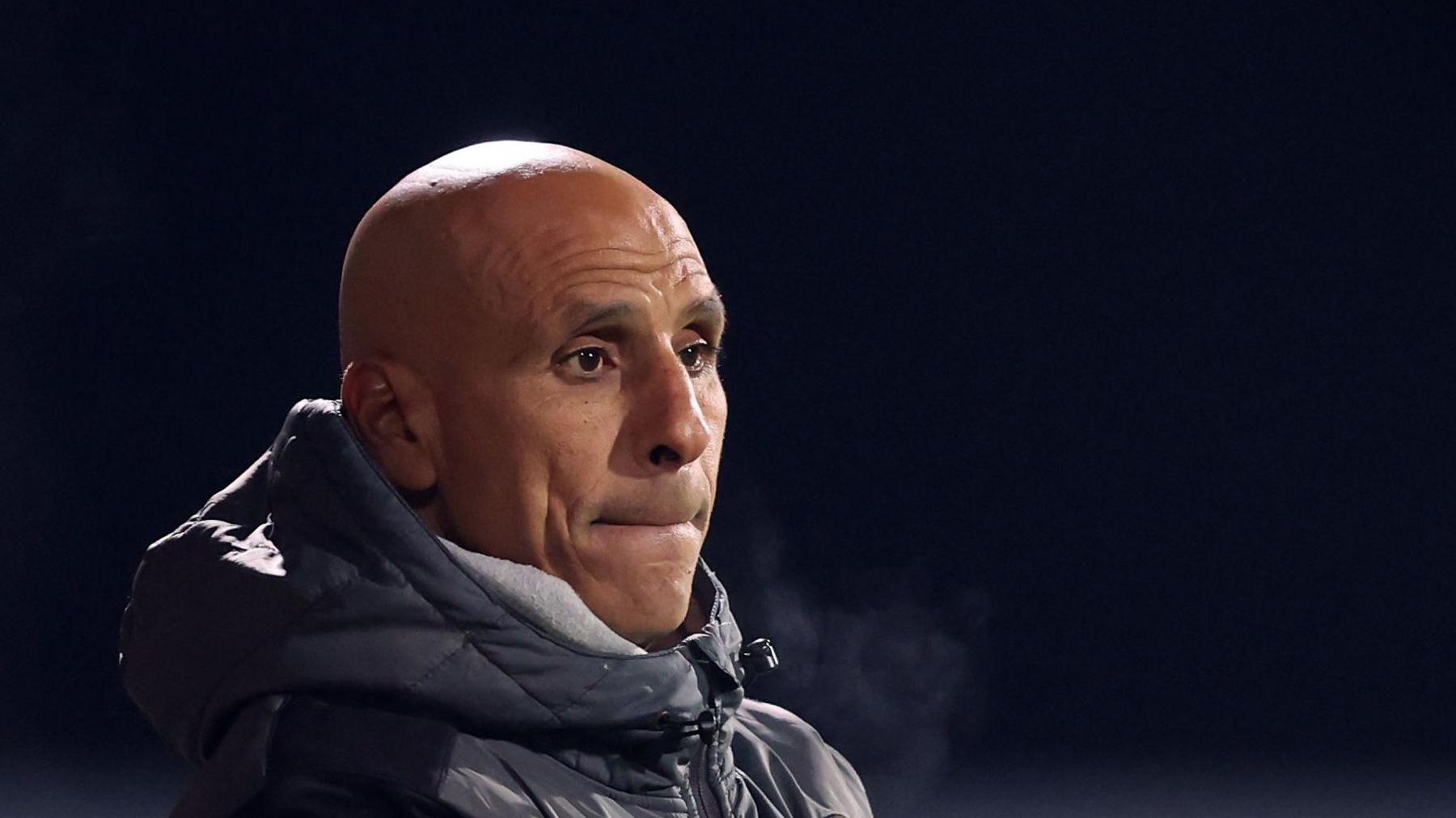 Dino Maamria watching a match with a big hooded puffa jacket zipped up to the top. His breath is visible