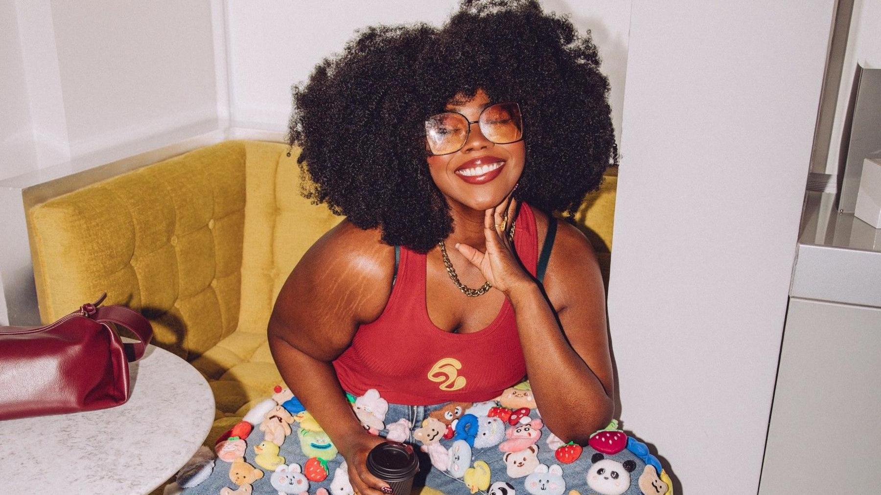 A woman wearing her natural afro hair is smiling at the camera. She's sitting on a yellow corner armchair, slightly leaned forward. She is wearing funky 70s inspired tinted glasses, a red top, and denim trousers with lots of little animals and fruits on!