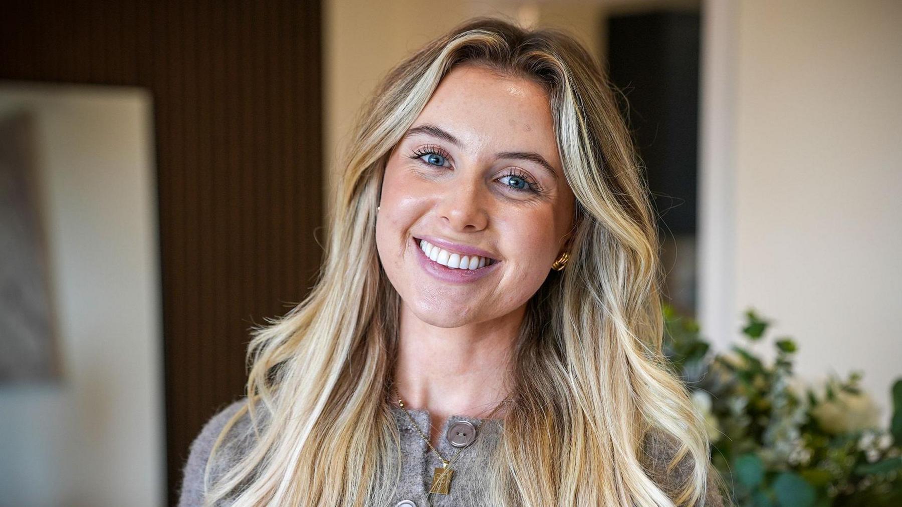 A young woman with blue eyes and long blonde hair looks into camera smiling, her head slightly tilted to the right. She is wearing a grey cardigan with big grey buttons and a gold necklace and earrings