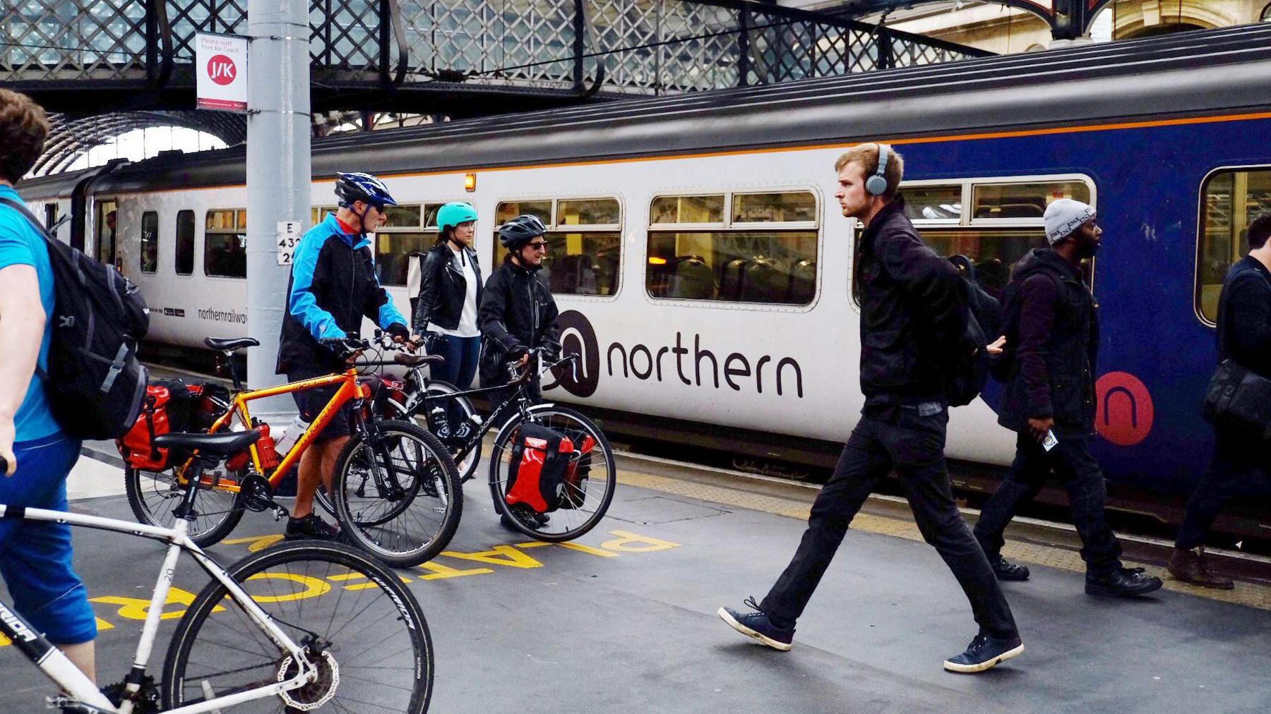 Commuters on a platform leave a Northern train on to a platform. The train is blue and white with the northern logo under the windows. The platform is busy with commuters wearing coats and bags and pushing bikes