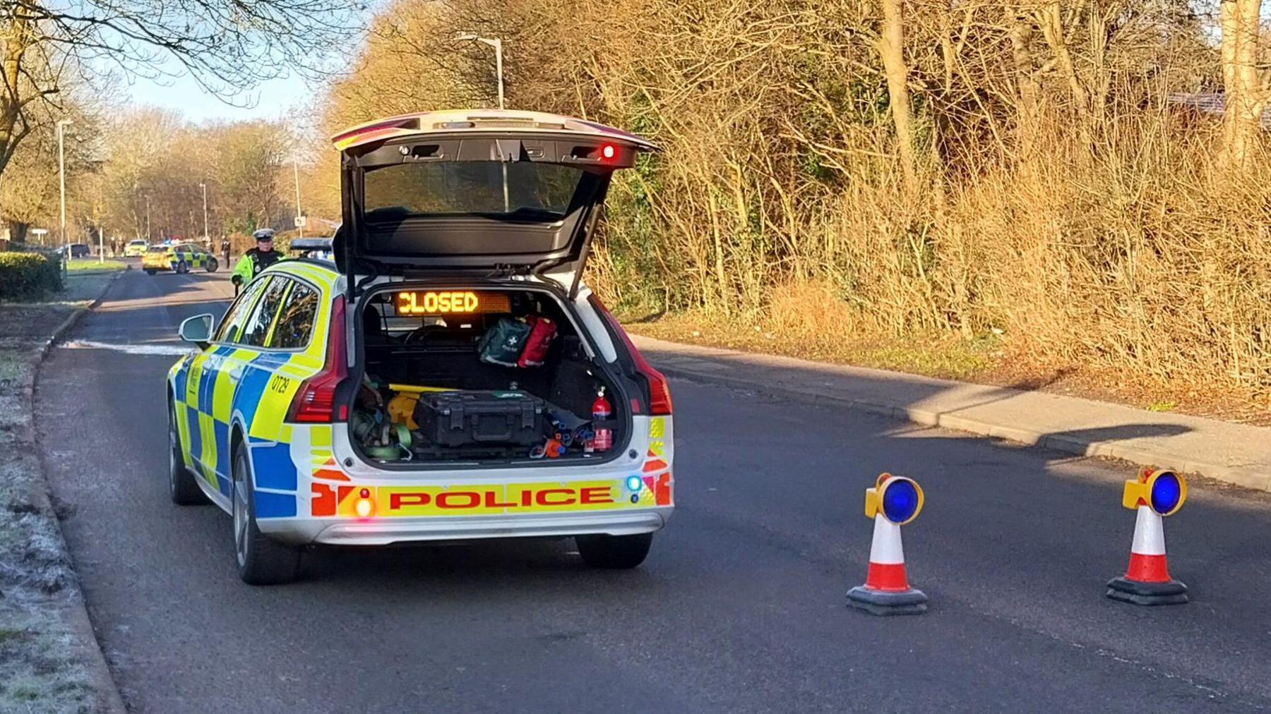 A police vehicle and cones blocking off a road. There is frost on the grass on a verge on on the left, and trees bathed in sunshine on the right. 