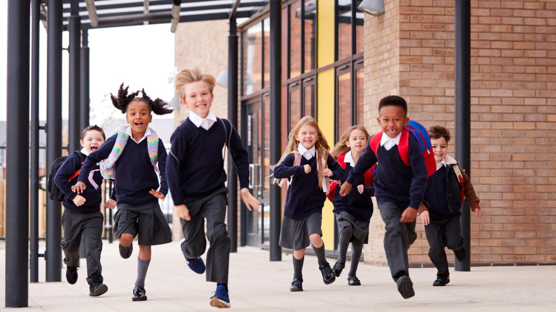 Primary school kids, wearing school uniforms and backpacks, running on a walkway outside their school building.