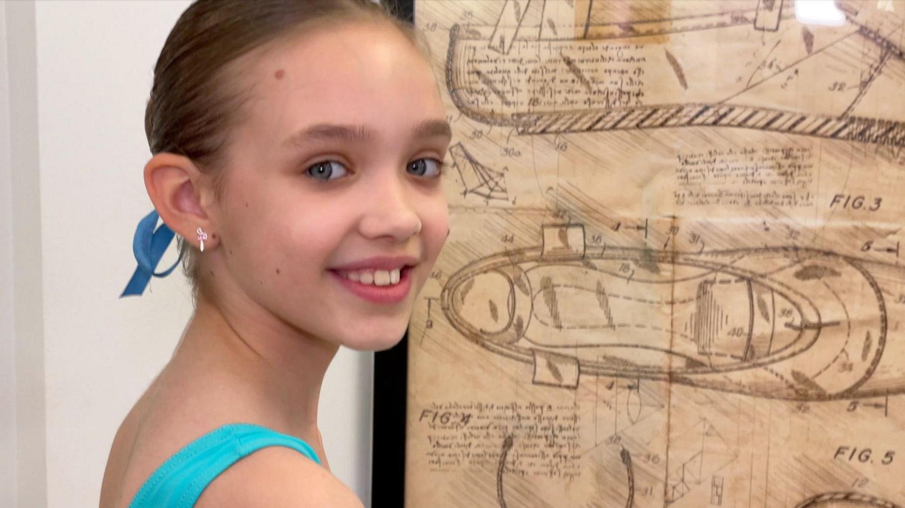 Jessica is wearing a blue leotard and is standing in front of a drawn diagram of a ballet shoe, which is stuck on a white wall. She is turn her head over her shoulder, smiling, looking at the camera.