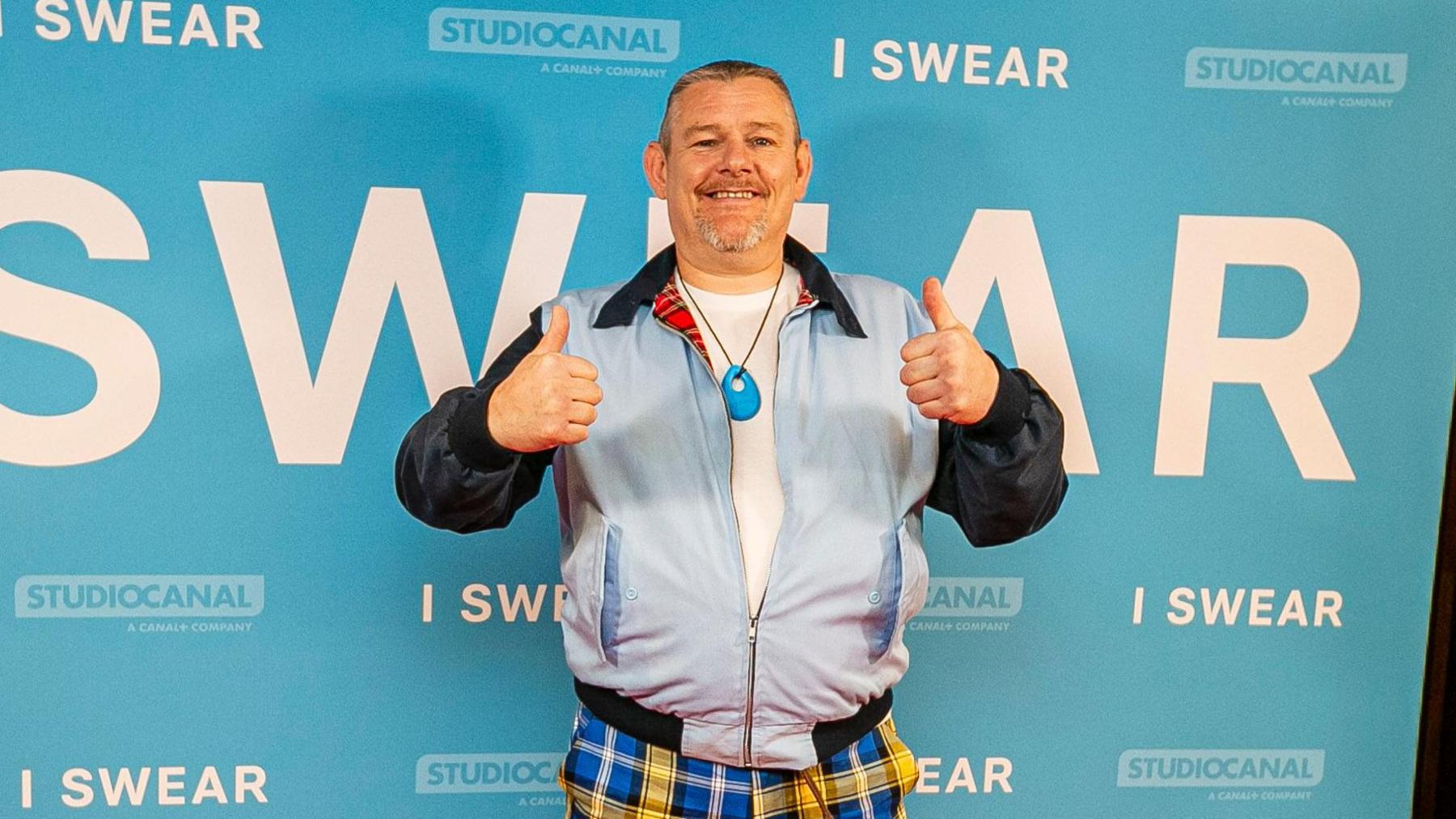 Photo shows John in front of a poster for the film I Swear. He's on a red carpet, wearing yellow and blue tartan trousers and a powder blue bomber jacket. He's giving a thumbs up gesture.