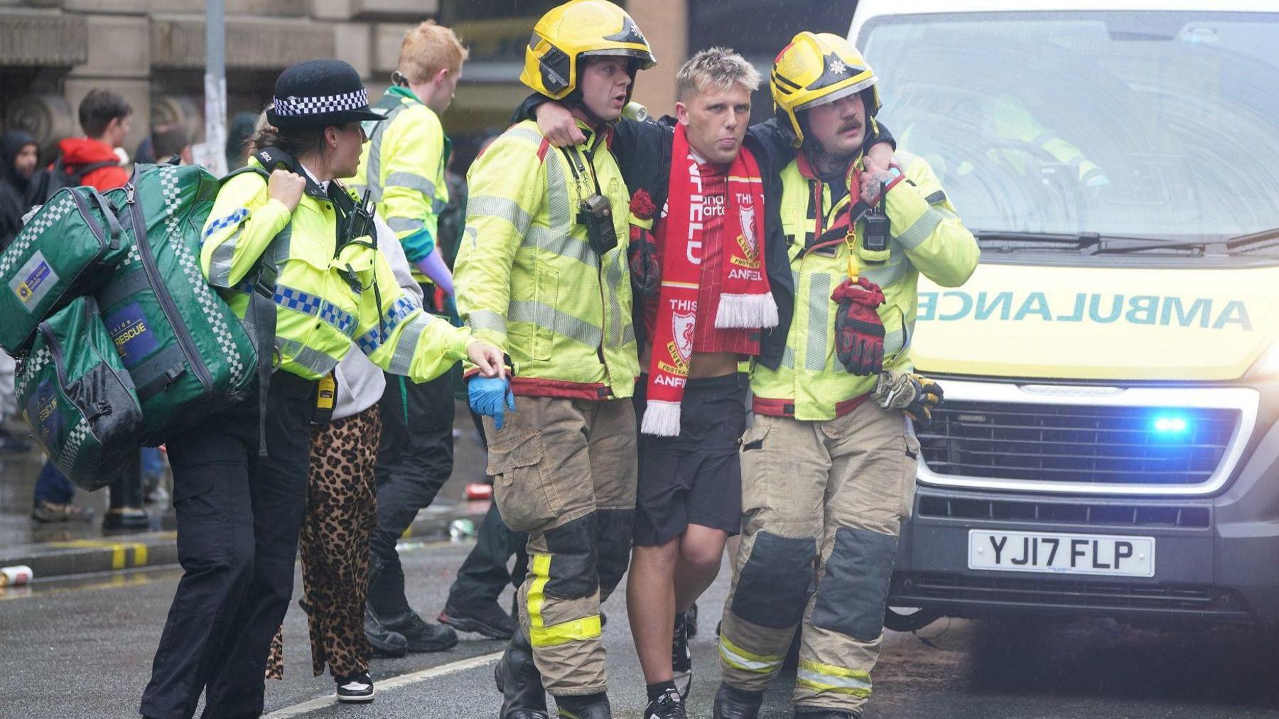 A young man with bleached blonde hair is wearing a Liverpool FC top and scarf with black shorts and trainers is being held up by two fire men who are wearing yellow reflective fire flak jackets, trousers and yellow hats. They are standing on a busy street and there is an ambulance in the background.