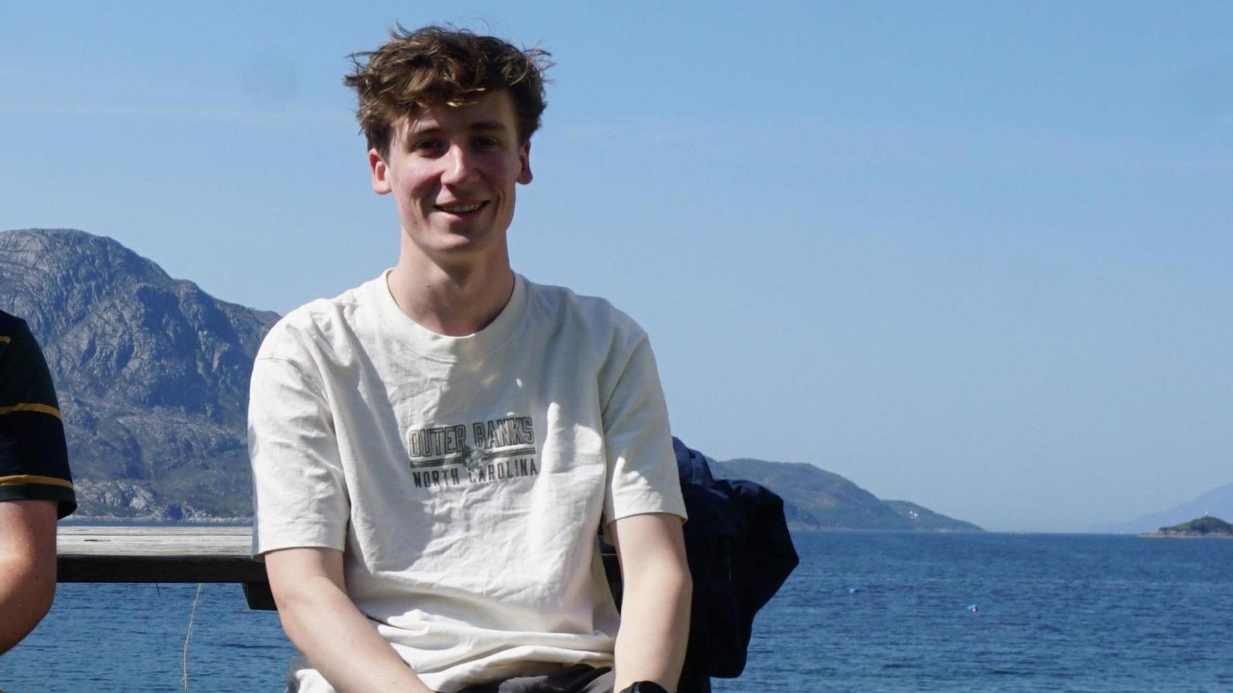 Caspar is sitting in the middle of the frame. He is wearing a white top. He is sitting in front of the sea and mountains