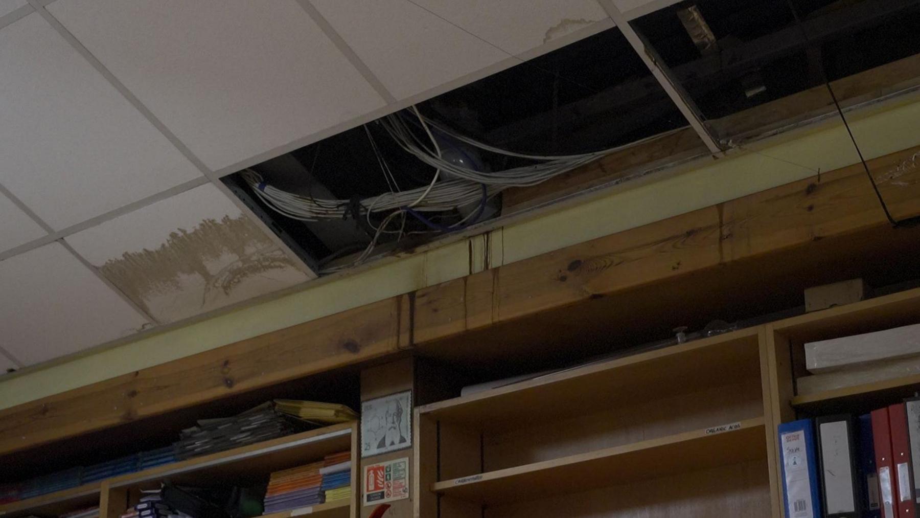 Ceiling tiles are missing revealing bunches of grey cable. Below this gap in the ceiling are shelves with books and binders.