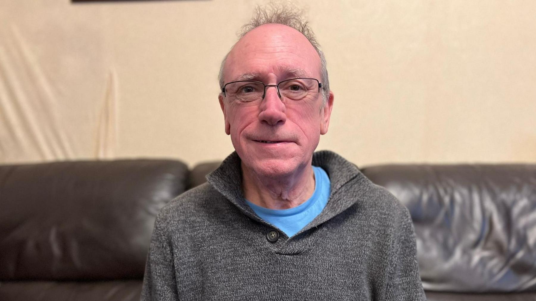 A man sits on a sofa. He has grey hair and glasses. He is wearing a grey jumper with a blue t-shirt underneath. The sofa is made of black leather and has a pink cushion on it.