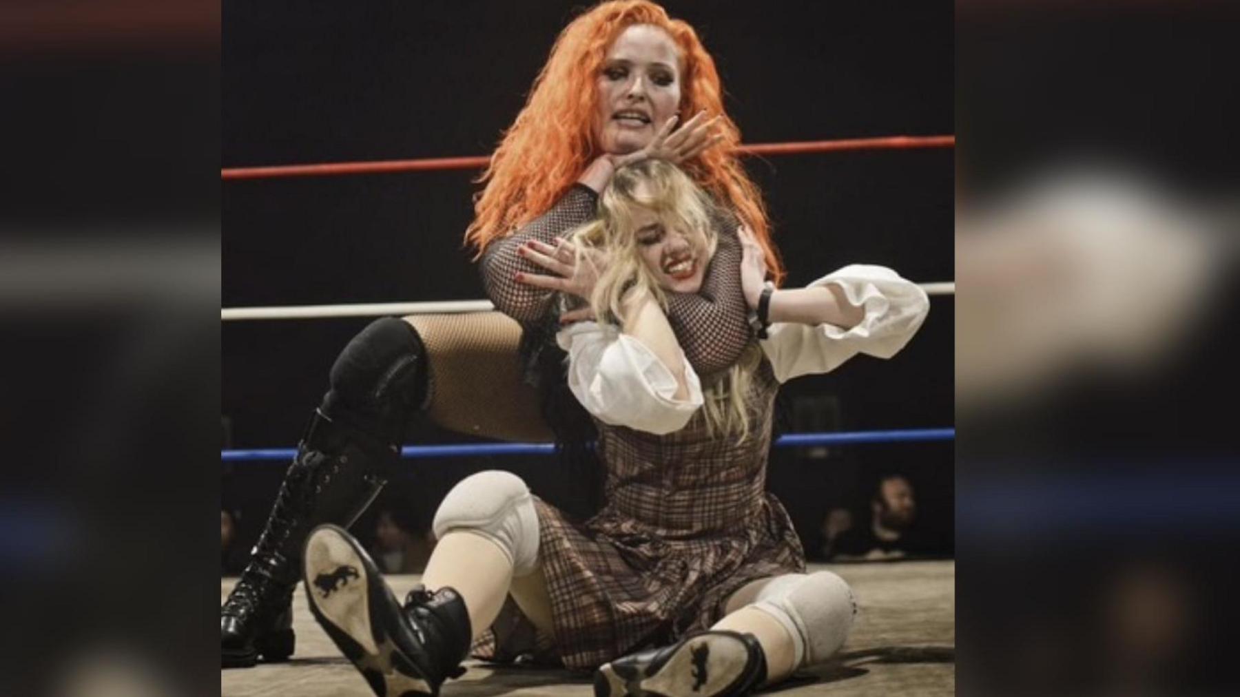 Annabelle at a wrestling match in a headlock by her opponent. Her opponent has bright orange curly hair and Annabelle is wearing a brown checkered dress.