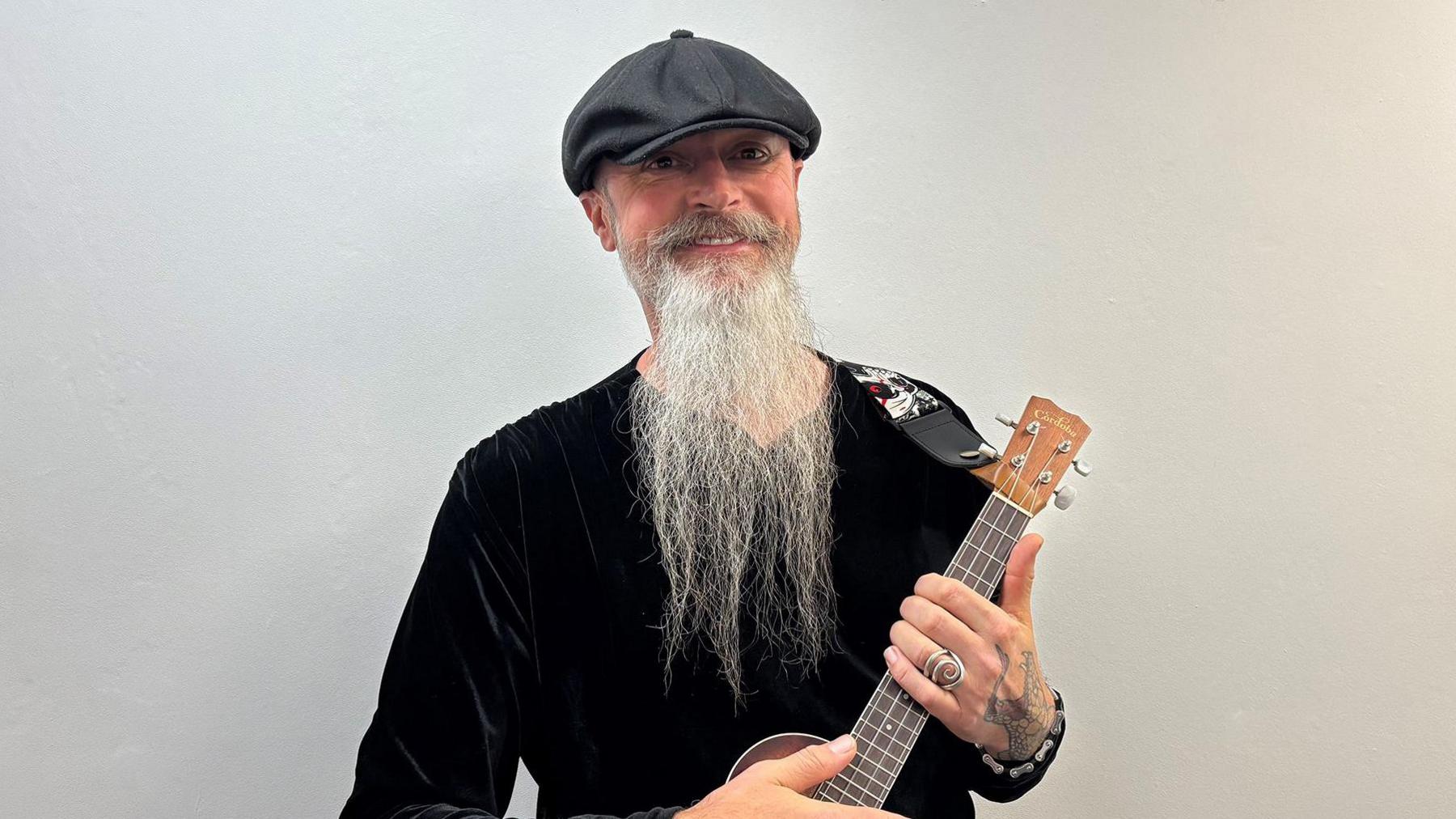 David Danby, a man with a long bear, is wearing a black top and black flat cap. He is holding a ukelele and is standing aganist a white backdrop.