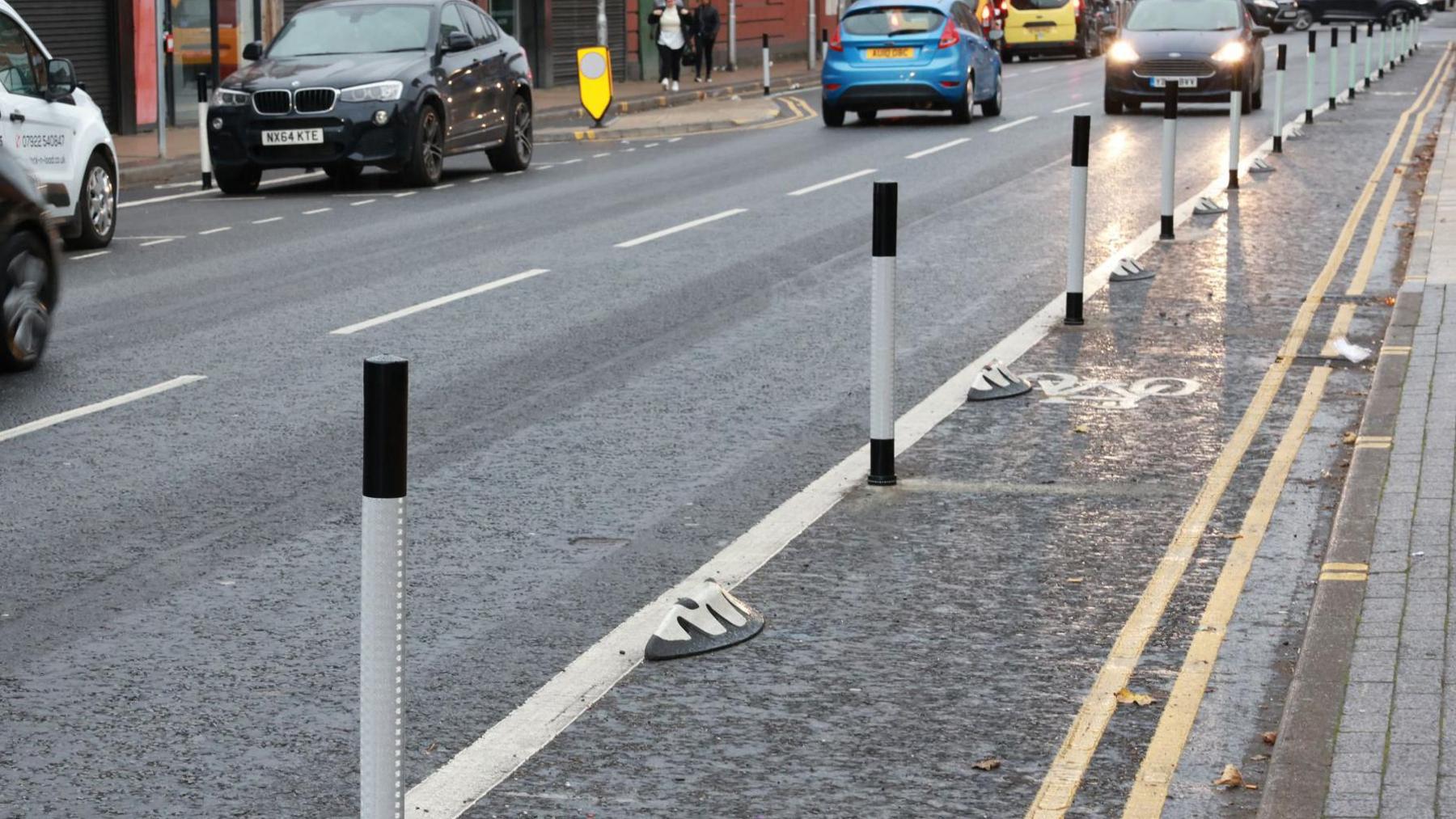 The cycle lane which is marked by a solid white line and black and white poles. Cars can be seen to the left of the cycle lane.