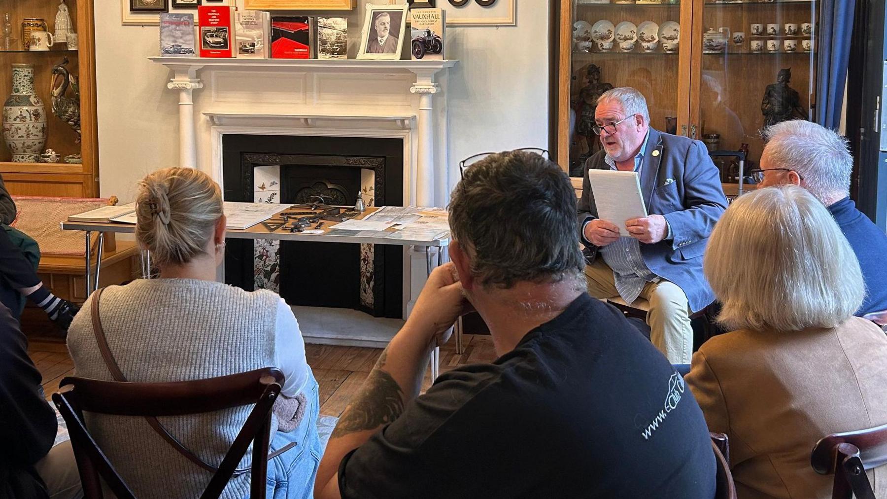 Andy King sits on a chair facing the camera and holding a pad of paper. His  mouth is open as he speaks to a small crowd of people seated in a circle, with their backs to us. There is a mix of younger and older people. To his right is a table with small vehicle parts, papers and photos. Behind him is a glass cabinet with museum artefacts.