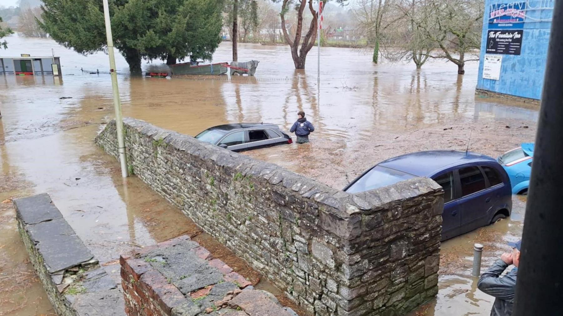 Deep flood water covers three parked cars. A man is standing beside one up to his waster in water. Another holds his hand to his mouth as he looked at stone wall walkway also flooded. 