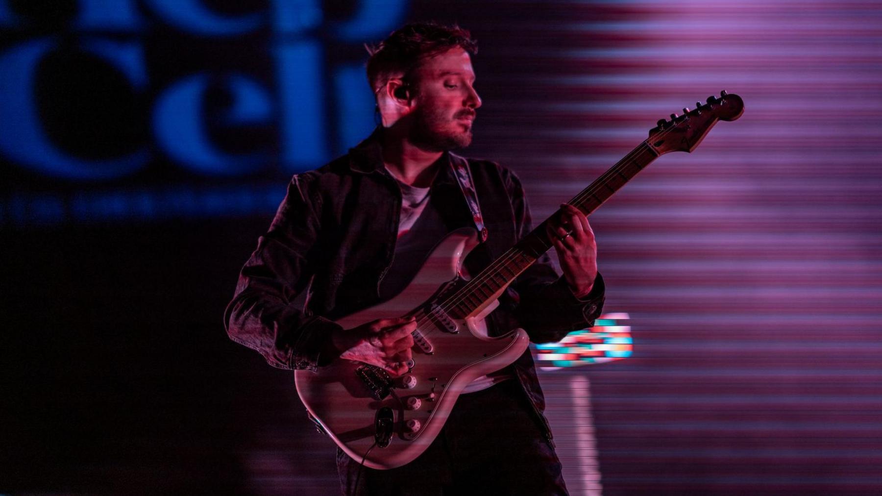 Tide Lines guitarist plays chords while strumming on his electric guitar. He has short hair and stubble on his face. The words "HebCelt" are just visible on a wall behind him in blue lettering. The guitarist is bathed in a purple/pink light.