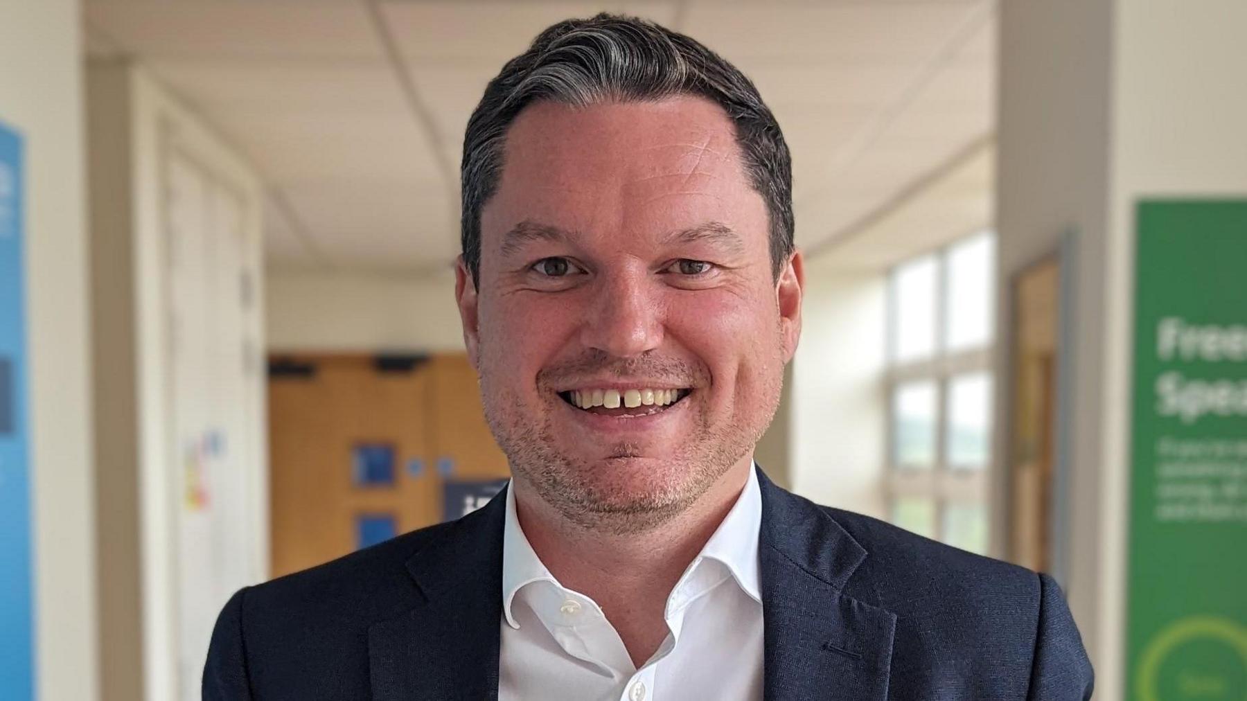 Hospital hallway in the background with Kevin McNamara in the foreground, smiling at the camera with stubble, wearing a dark blue blazer and white shirt