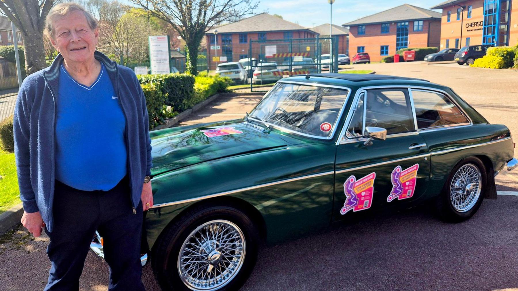 Dave Whittington wearing a blue t-shirt and cardigan, standing near the bonnet of his dark green 57-year-old MGC classic car.