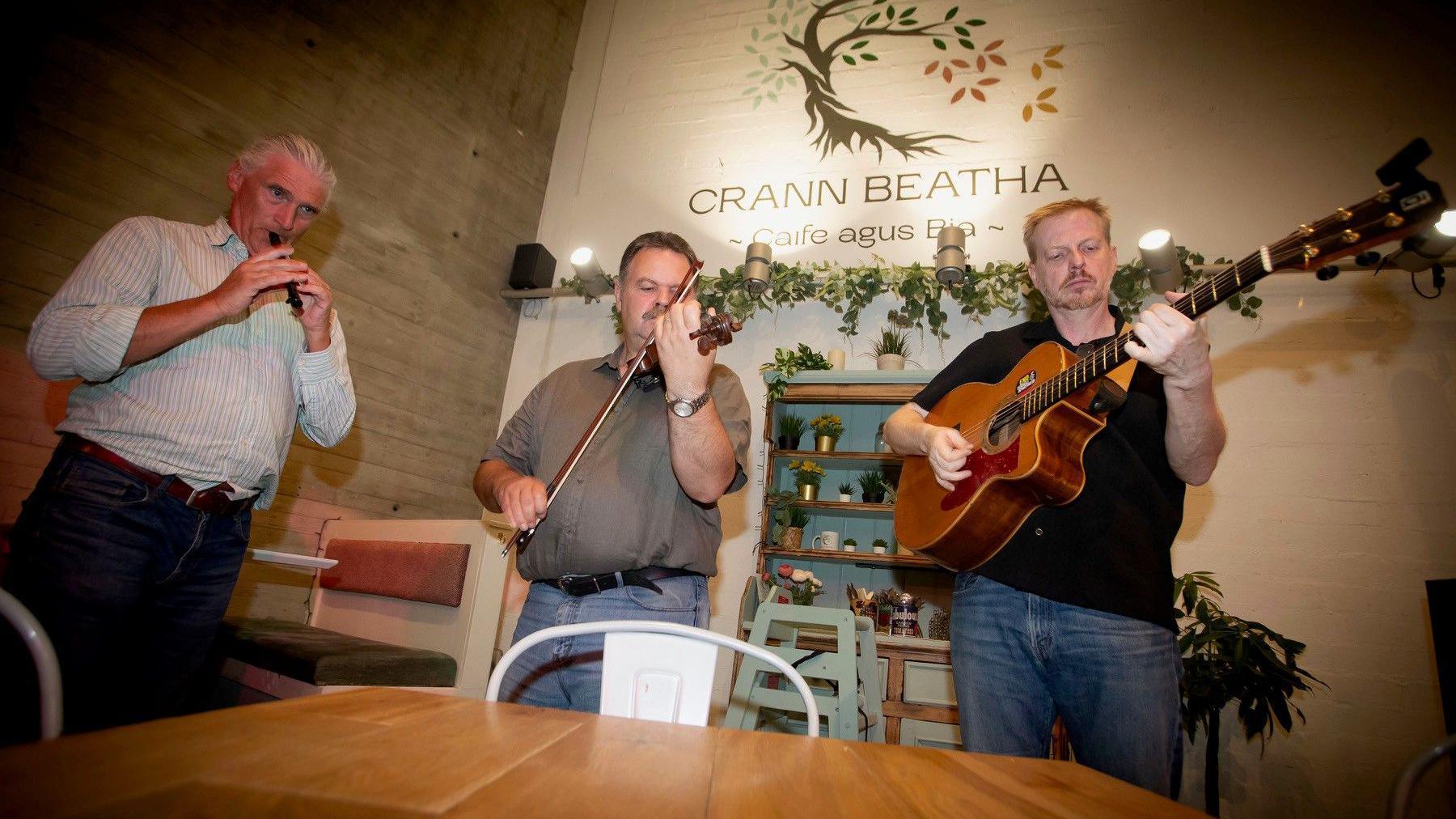 Three men are playing instruments of a recorder, violin and a guitar. On the wall it says 'Crann Beatha'.  
