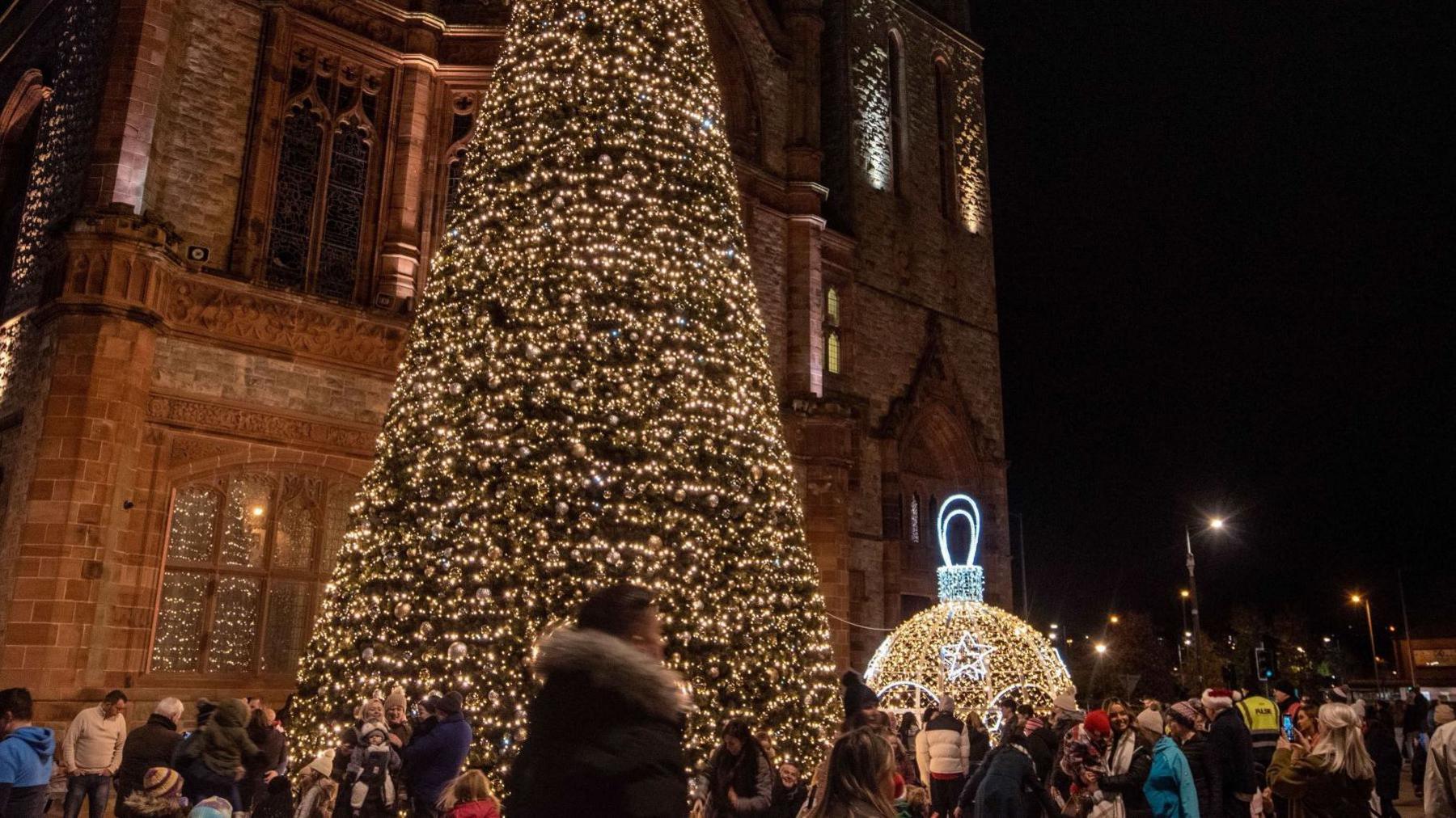 A large crowd look up at the lit Christmas tree outside the Guildhall in Derry. It is a large stone building.