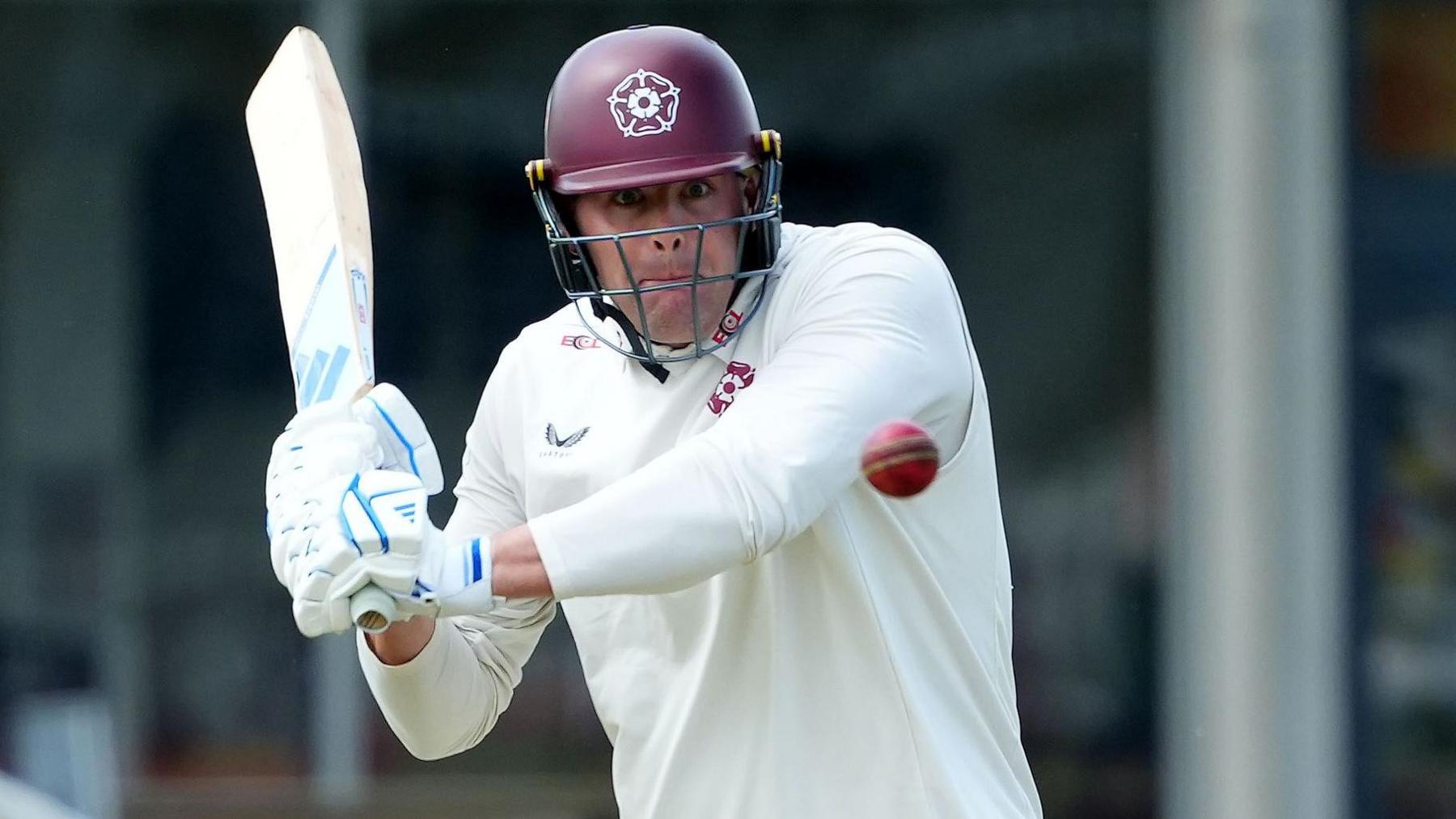 Louis Kimber in a maroon helmet and white shirt swings his bat towards the ball