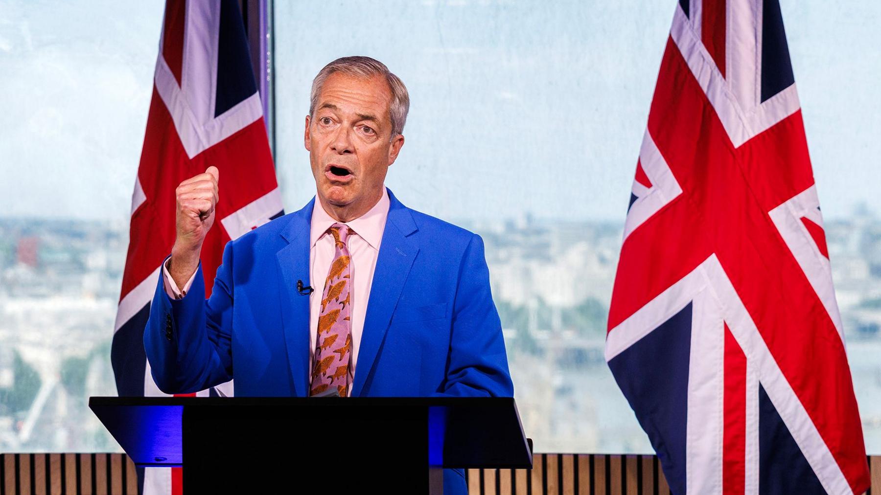 Nigel Farage in a blue suit and pink shirt and tie, standing in front of a blue background with a union flag in the background