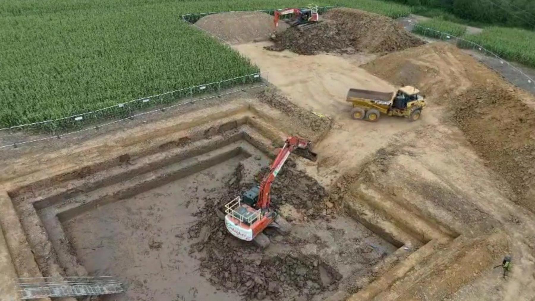 A mechanical excavator digs up a field. A dumper truck is parked nearby.