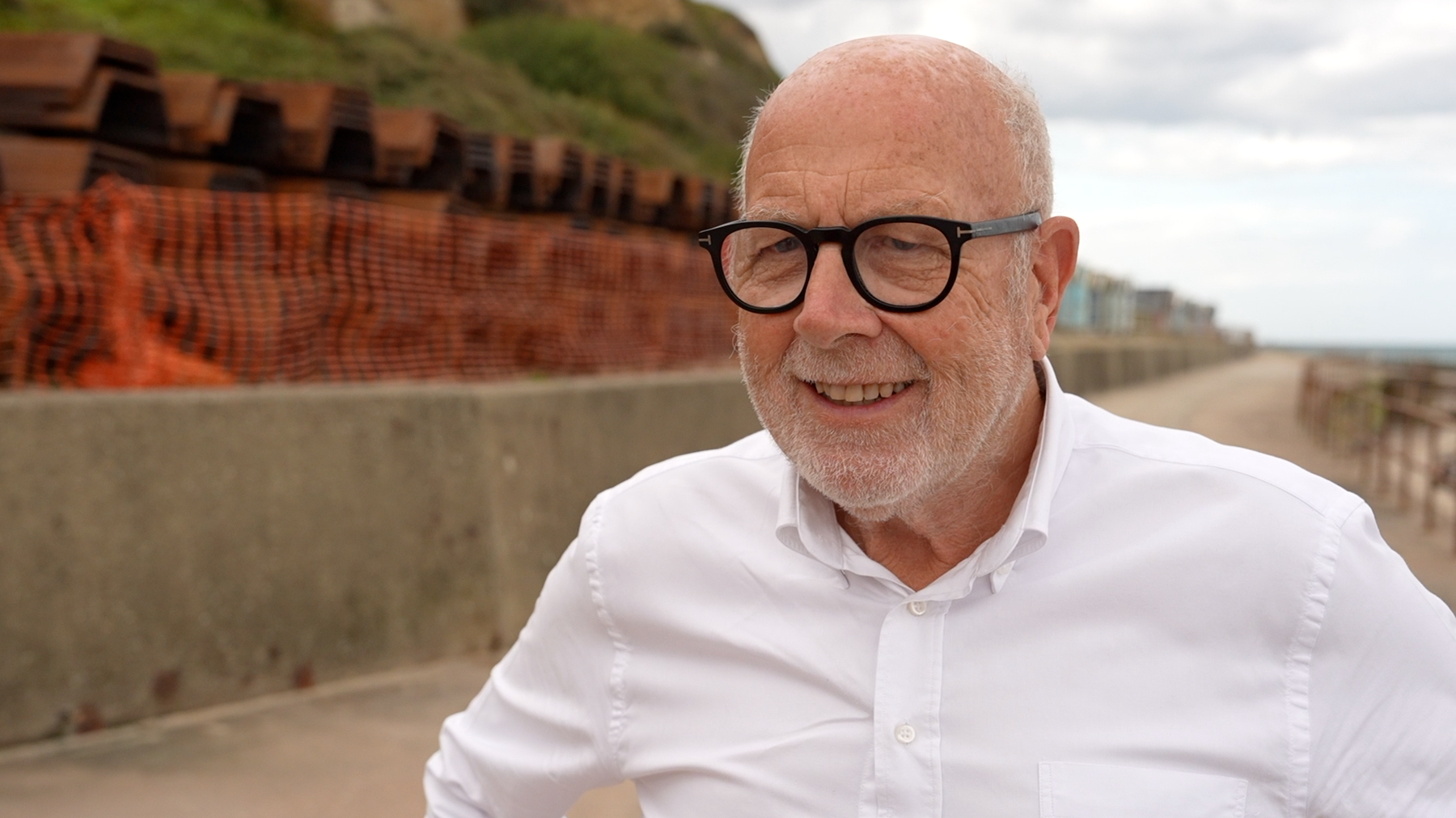 Harry Blathwayt standing outside on the seafront in Overstrand. Behind him are steel panels laid out ready to be used to reinforce the sea wall - they are cordoned off from the public with a row of orange netting. Blathwayt is looking to the left of the camera and is wearing a white shirt and a pair of black circular framed glasses on his face.