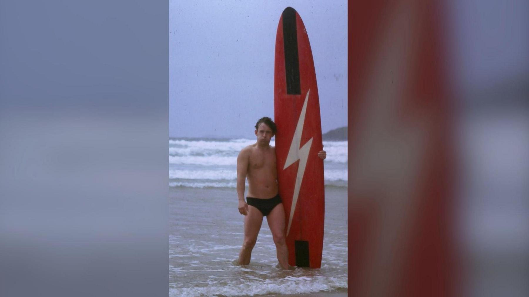 A young man average height in black swim trunks holding a red surf board with a white lightning bolt. The board is twice his size. 