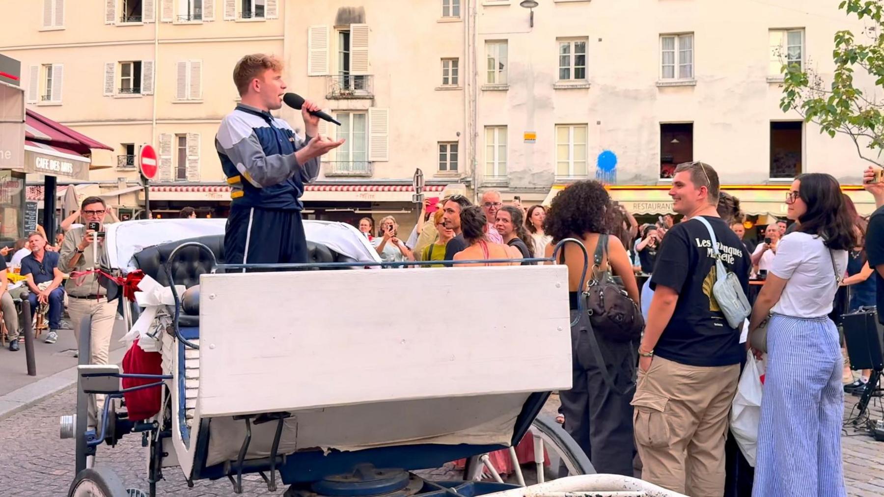 Mickey Callisto dressed in a navy blue track suit while singing. He is standing in a white carriage surrounded by surprised on lookers in the Place de la Contrescarpe in Paris.