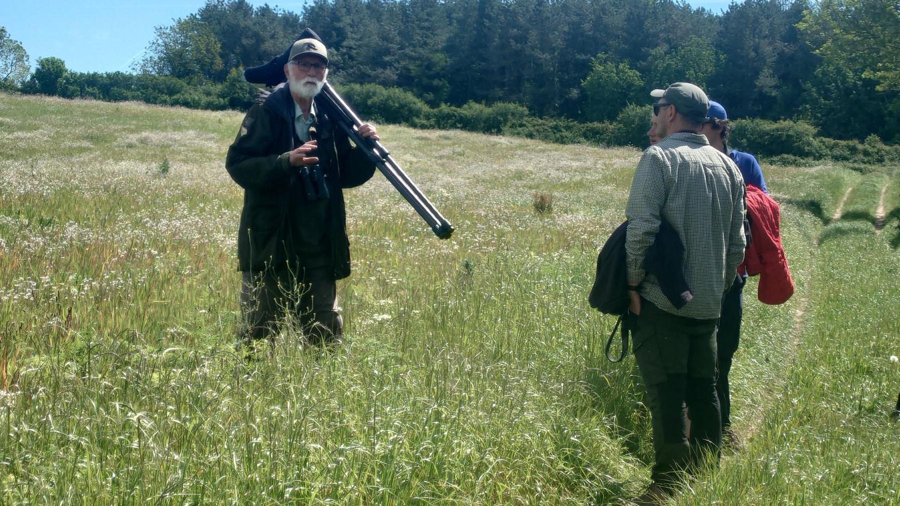 Nigel is standing in a field of tall grass and talking to several people standing on a grassy path. There are trees in the background. 