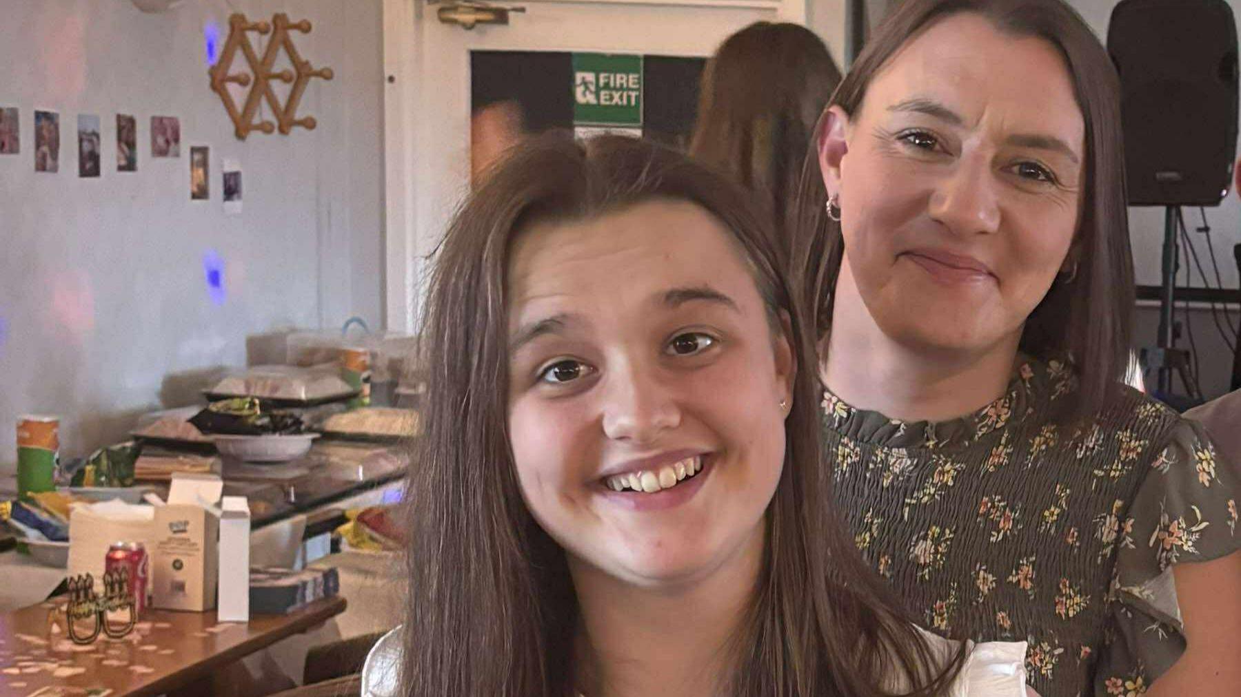 Emily with long brown hair wearing a white top standing in front of her mother Victoria with long brown hair wearing a black and flowered patterned blouse at an event with buffet food in the background. They are both smiling.