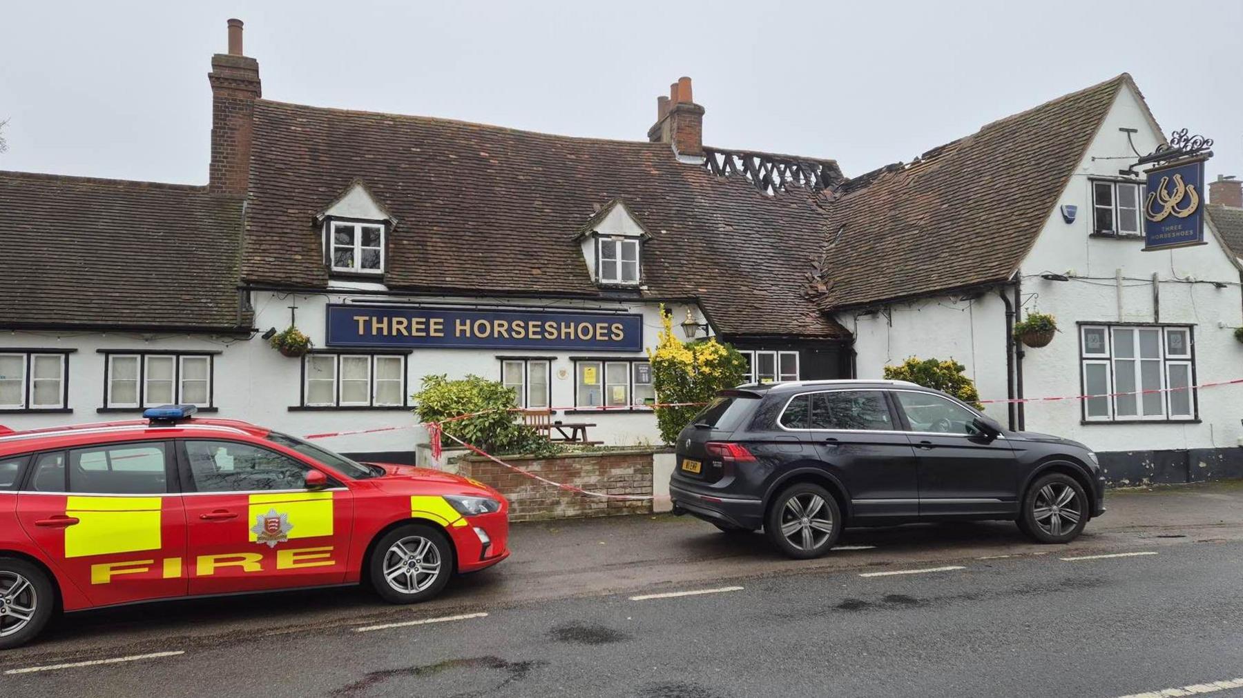 Two cars are parked on the pavement outside the pub. One is a red-and-yellow fire service vehicle and the other is dark blue or black. There is tape cordoning off the entrance to the pub. The fire-damaged beams in the roof can also be seen.