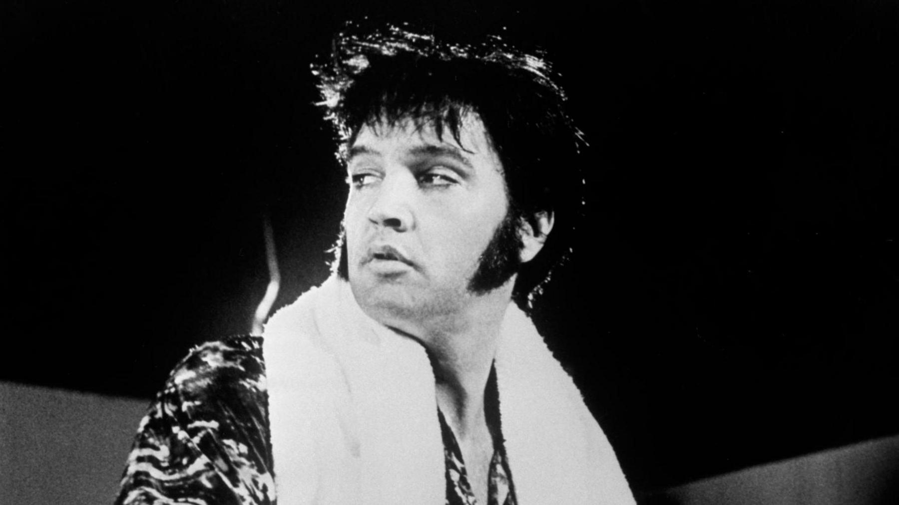 A black and white image of Elvis Presley, who is wearing a shirt and has a white towel wrapped around his shoulders.