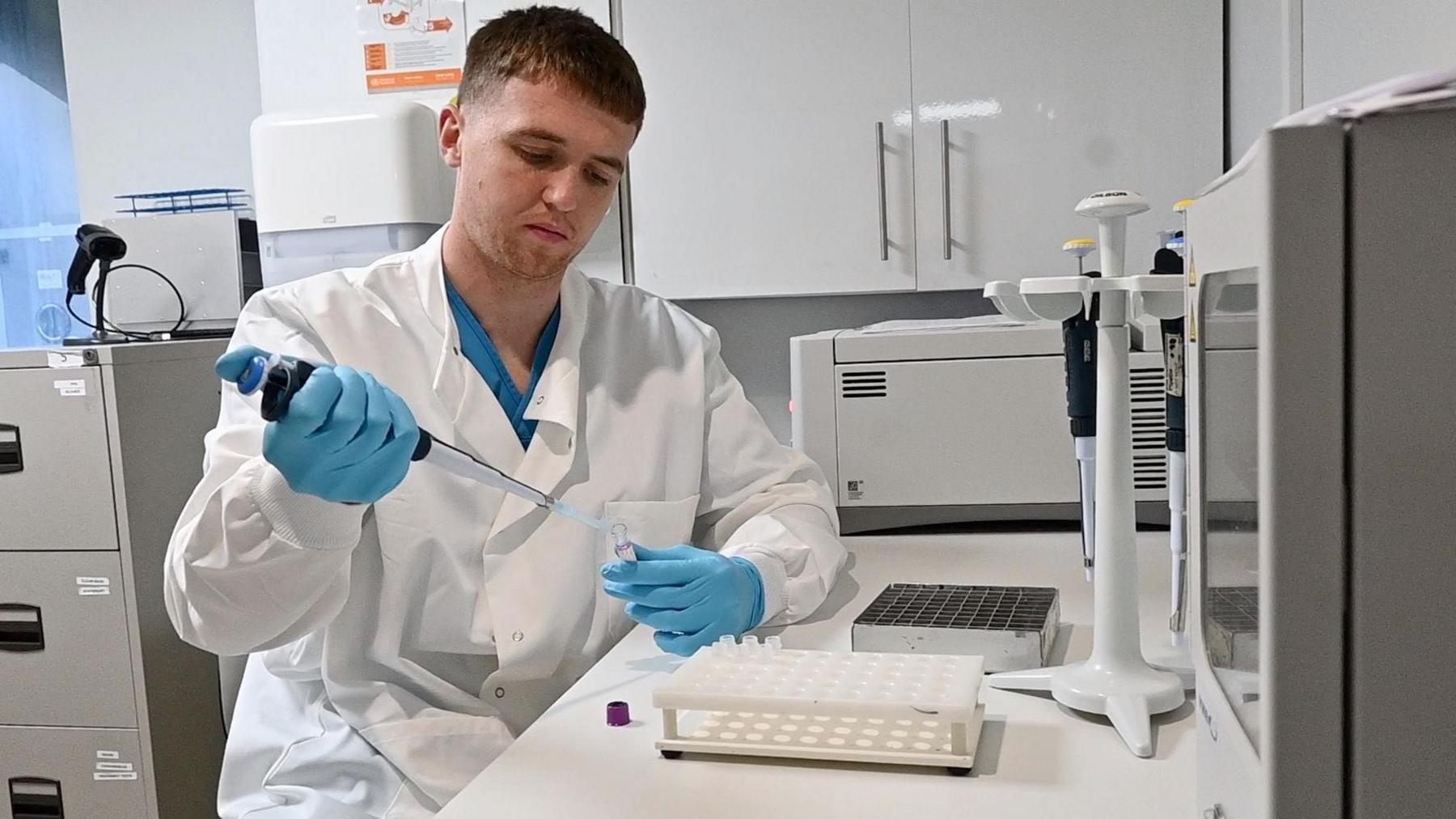 A man wearing blue gloves and a white, lab coat using a large pipette to insert a substance in a small tube. He appears to be in a white greyish lab.