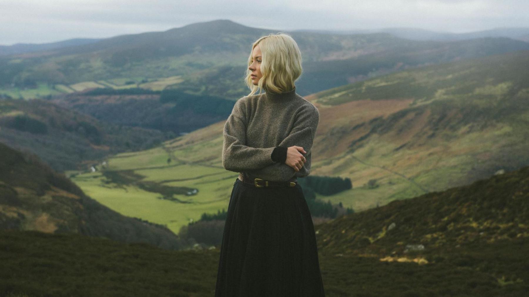 Nina Nesbitt is standing with her arms folded. She is looking off into the distance and is pictured in a rural landscape.