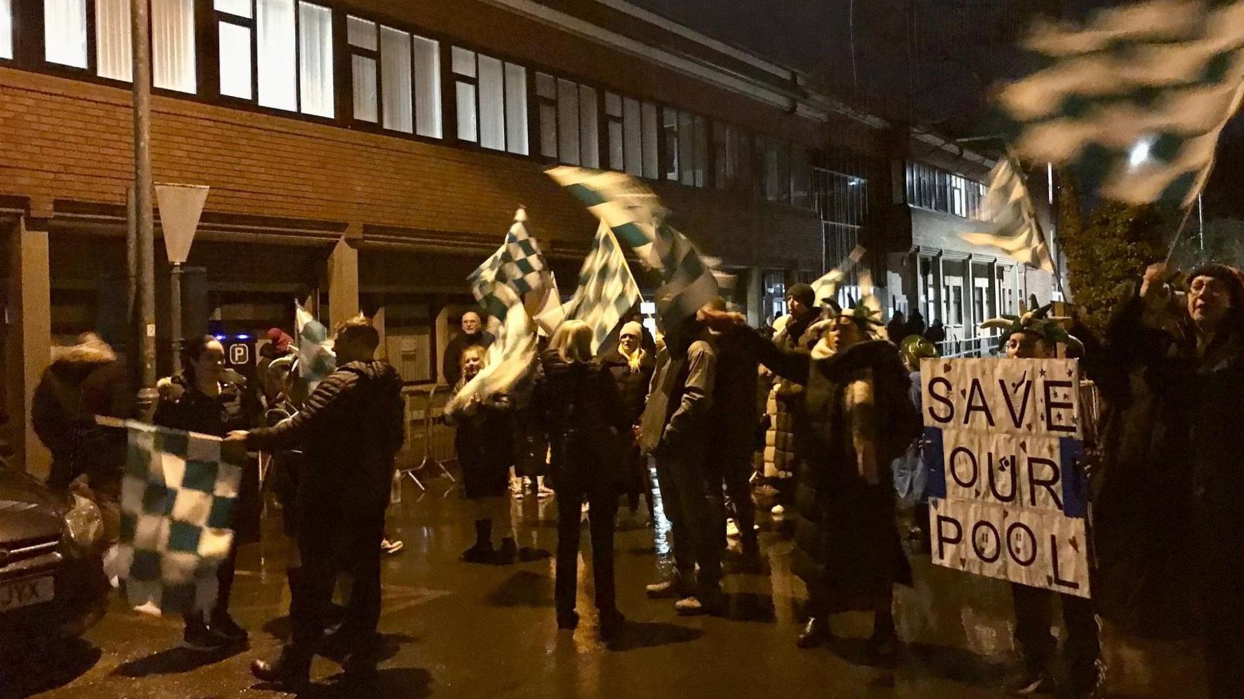 Protesters waving flags and holding banners gather in the dark outside the meeting.