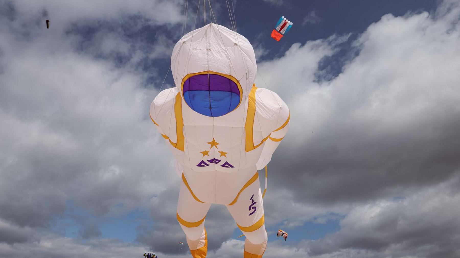 A large spaceman kite flying high above the beach. It has a white suit with yellow trim and a purple face mask.