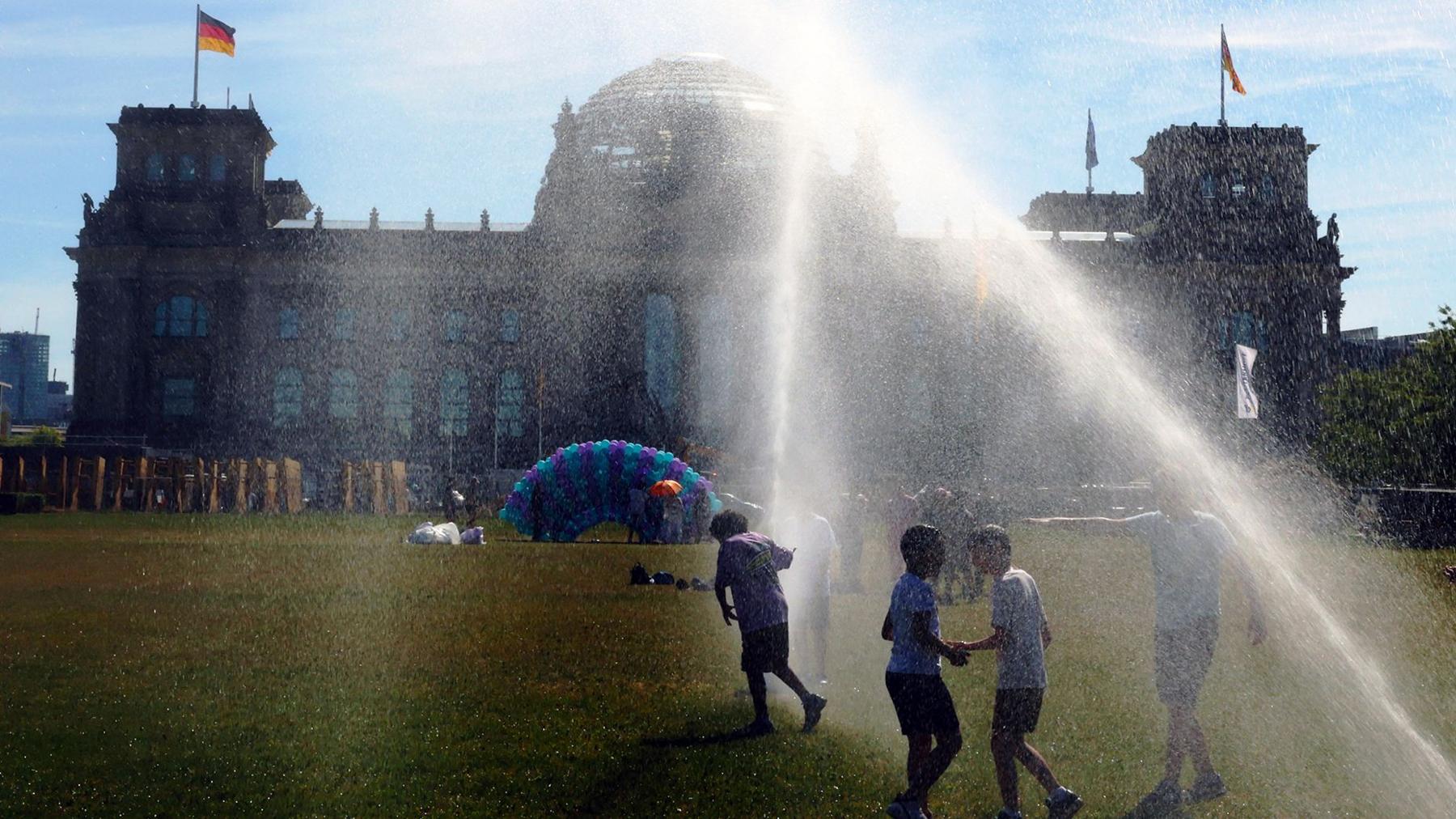 People pass by a watering system in front of the Reichstag on a hot summer day on June 24, 2026 