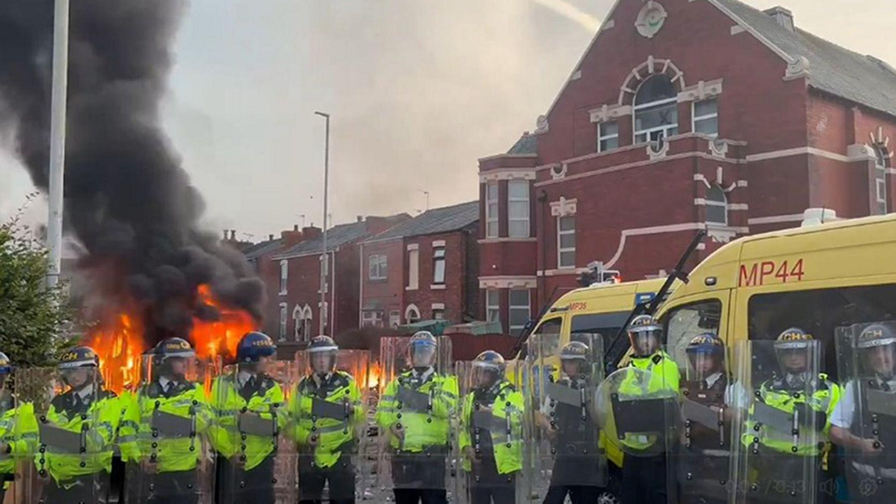 A police van on fire and riot police officers in a line in front of mosque in Southport