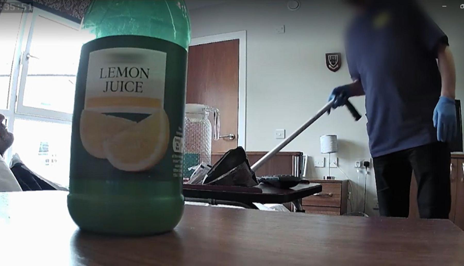 A grab from Susan's secret filming footage in the care home room. In the foreground is a bottle of lemon juice which has been placed on a desk in fronyt of the secret camera. In the background is a cleaner holding a walking stick. A figure on a bed is also visible.