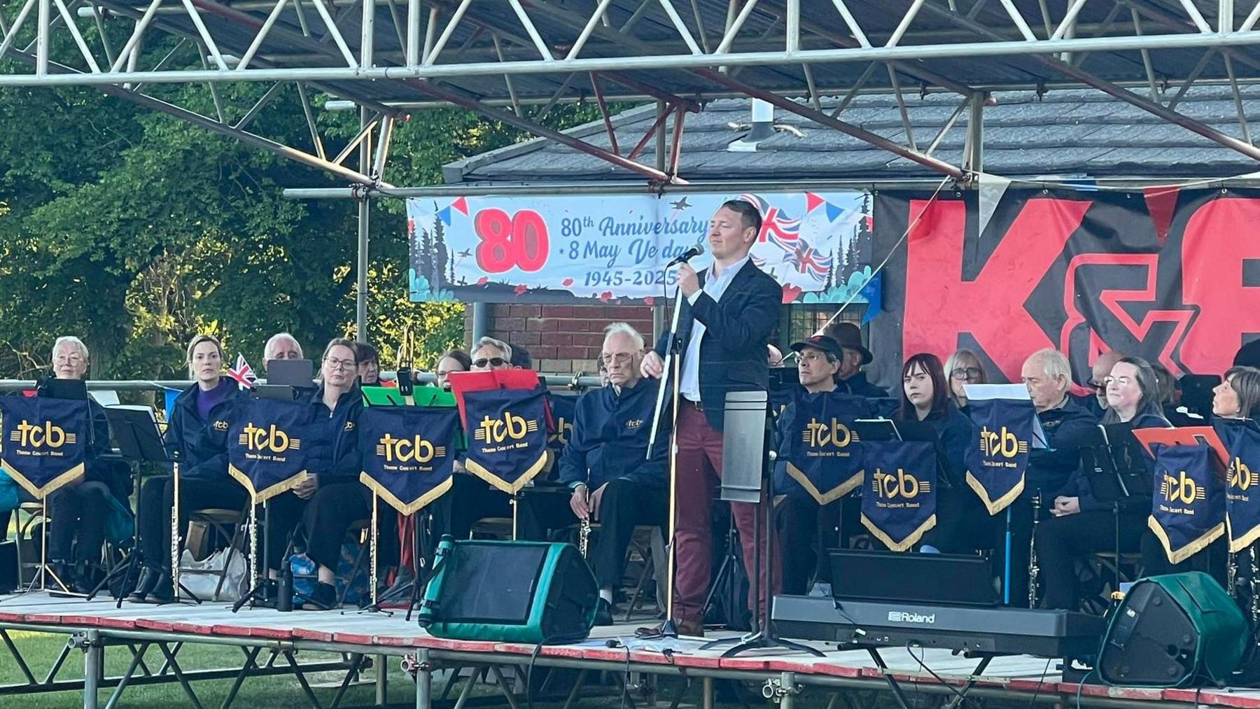 A big band on an outdoor stage with a man standing in front at the microphone