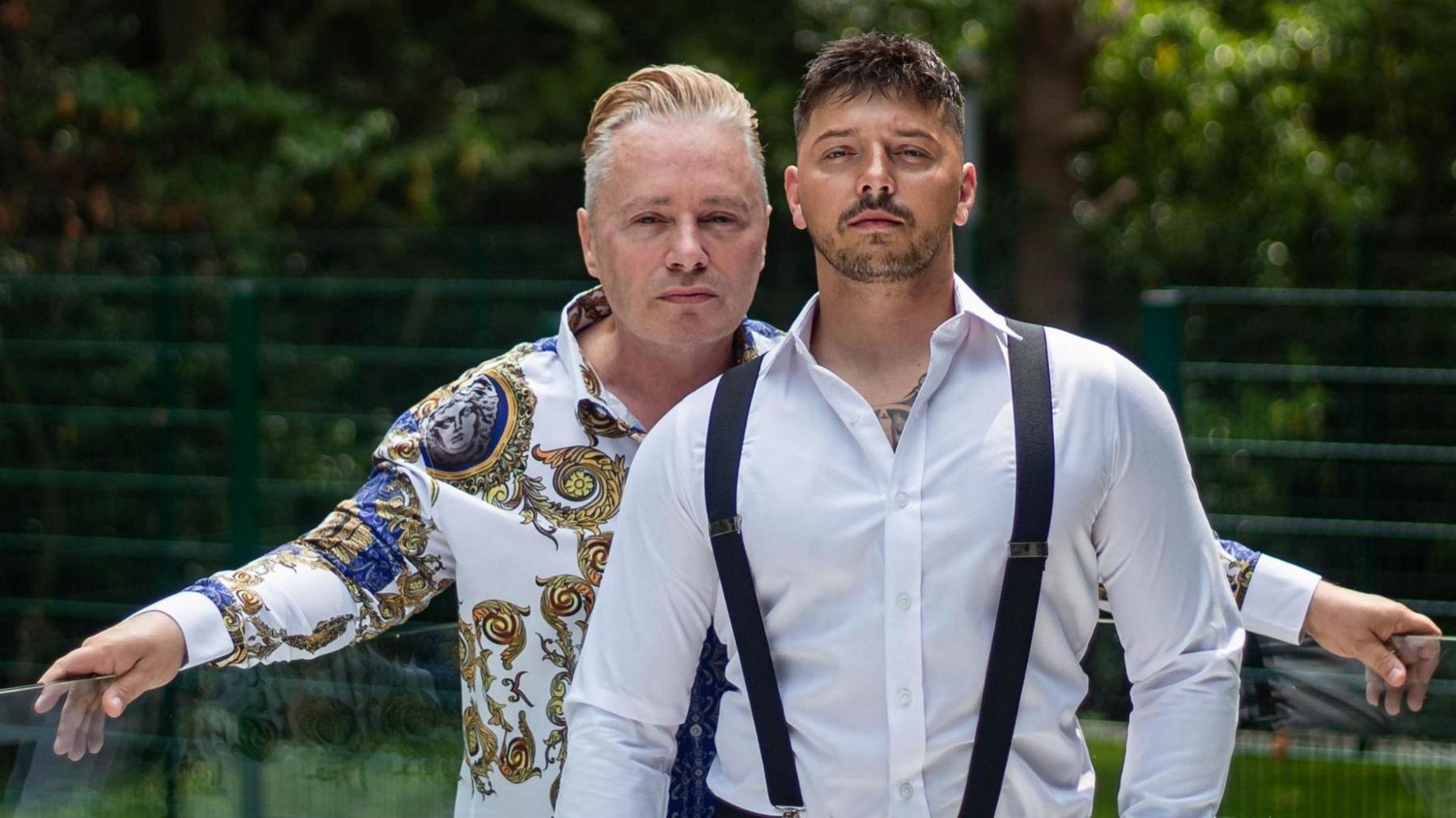 Barrie and Scott Drewitt-Barlow standing on a glass balcony, with trees and a fence in the background. Barrie is wearing a white shirt with gold patterns on it. Scott is standing in front of Barrie, wearing a white shirt with black braces. They are both looking at the camera with a serious expression