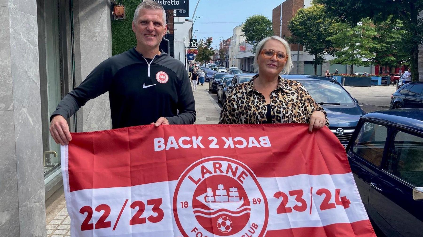 A man and a woman stand on the street in Larne. The man is wearing a black tracksuit top with a Larne crest, and the woman is wearing a leopard print shirt. The man has short grey hair and the woman has short blonde hair and glasses. They are holding a flag: the flag says ‘Back 2 Back Champions, 22/23, 23/24’ with the Larne crest in the middle. The Larne crest is a white circle with a red middle. In the middle is a white sail boat.