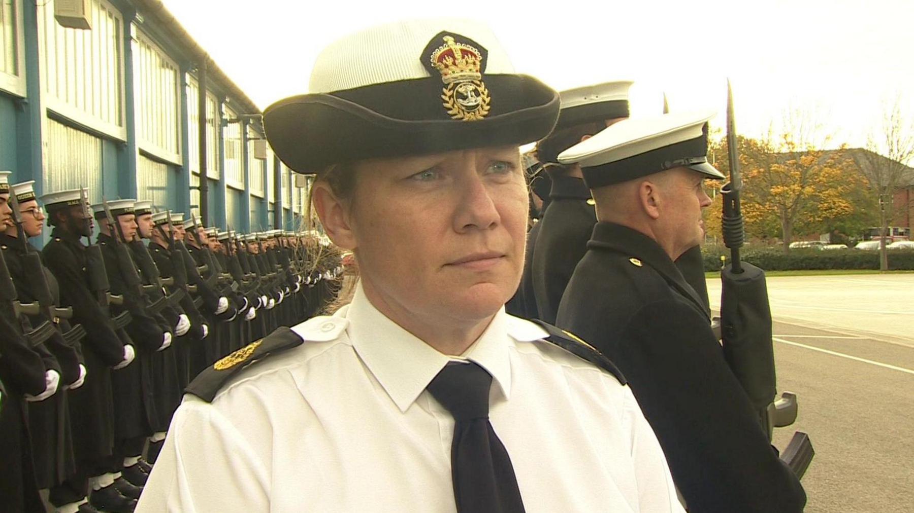 CPO Evans wears a cap, white shirt and black tie. Armed personnel are stood to attention behind her.