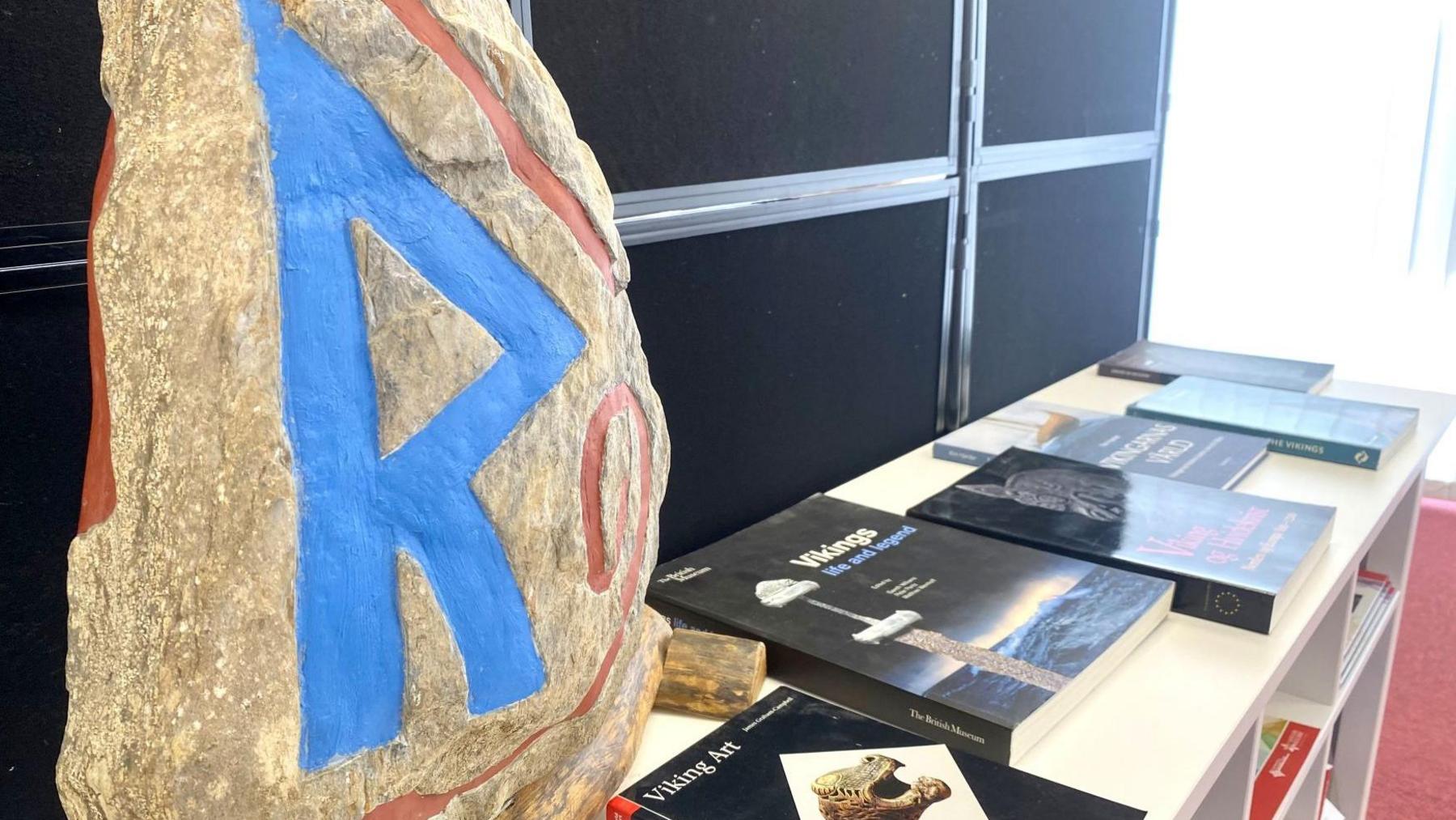 A large rock with the letter R painted on it in blue, on a white table alongside a number of history books about Vikings.