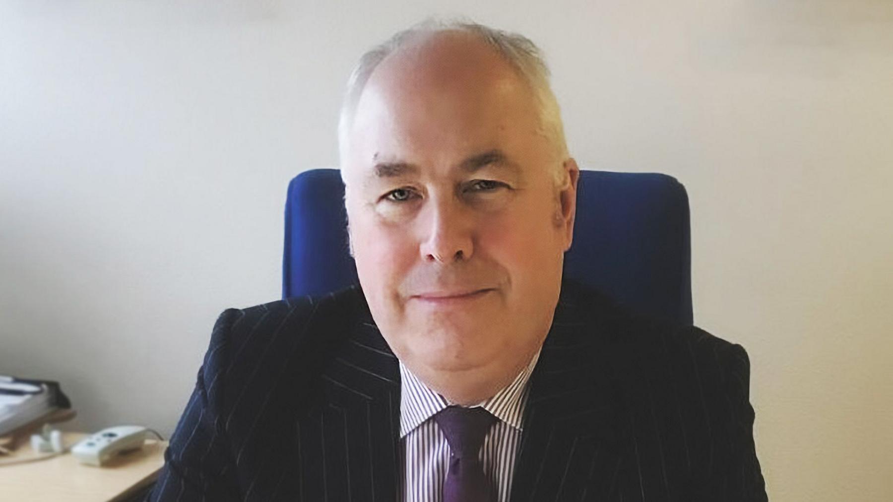 Malcolm Burr - a man with grey hair pictured sitting at his desk smiling wearing a pinstripe suit and purple tie