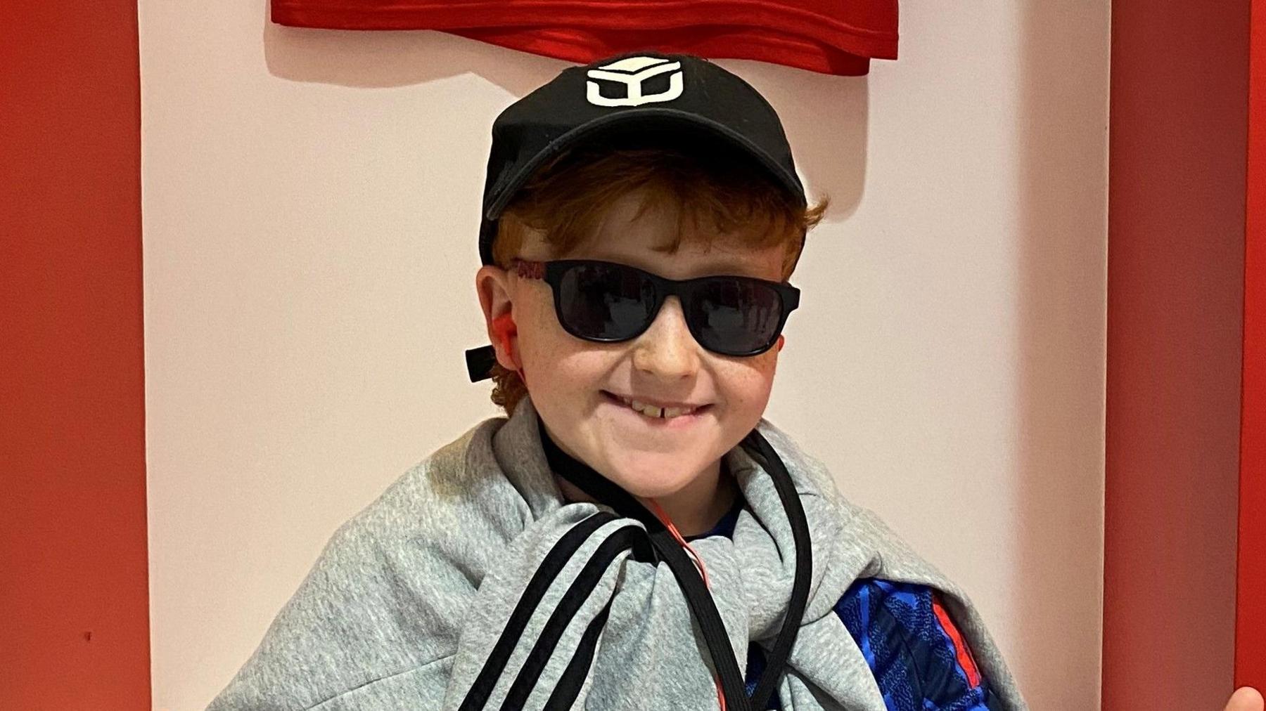 A young boy sits in a football changing room smiling at the camera. He has both thumbs raised up. He wears a black cap and sunglasses and has a grey jumper tied around his neck.